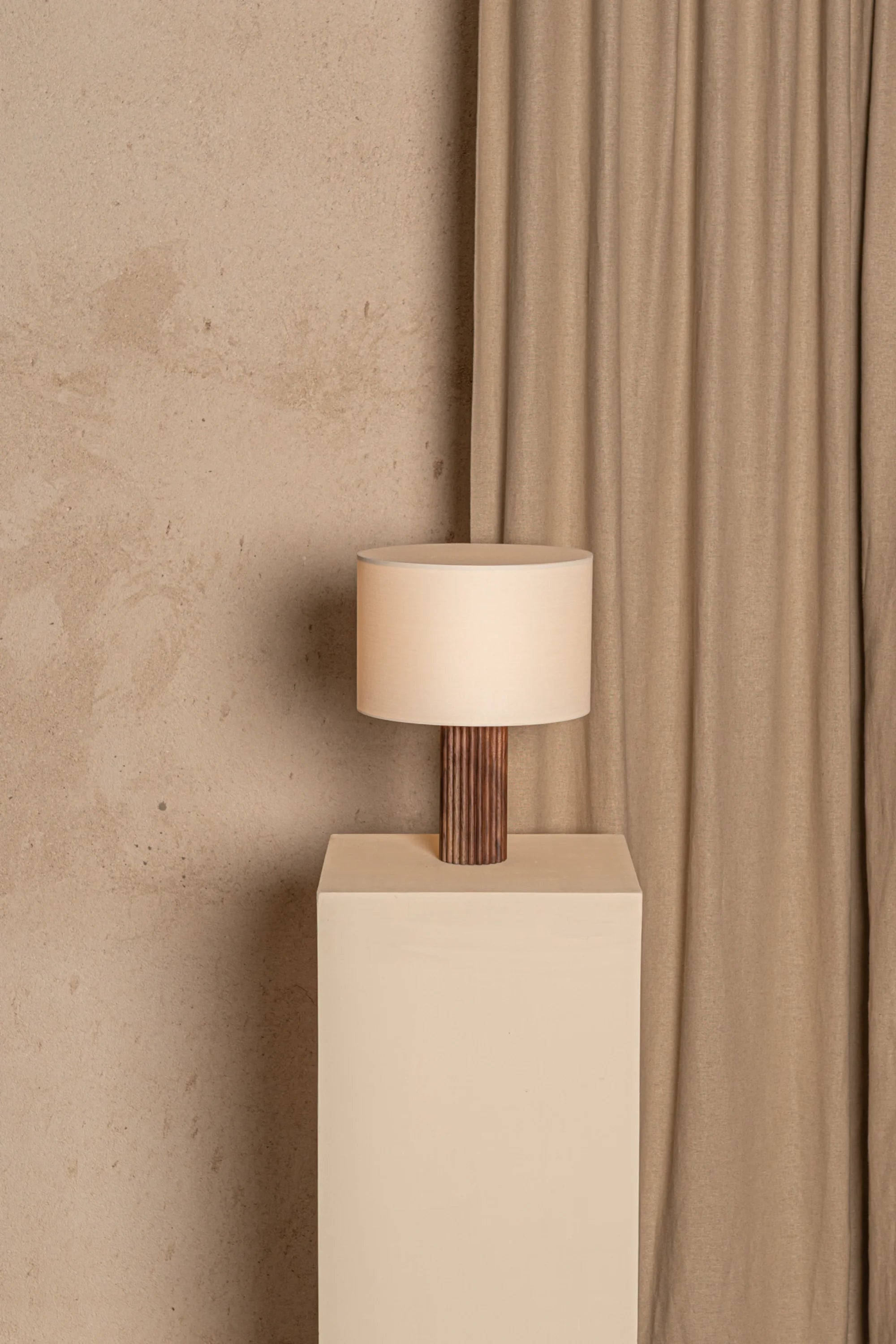 Table lamp with wooden base and beige shade on a white pedestal against a beige wall and curtain.
