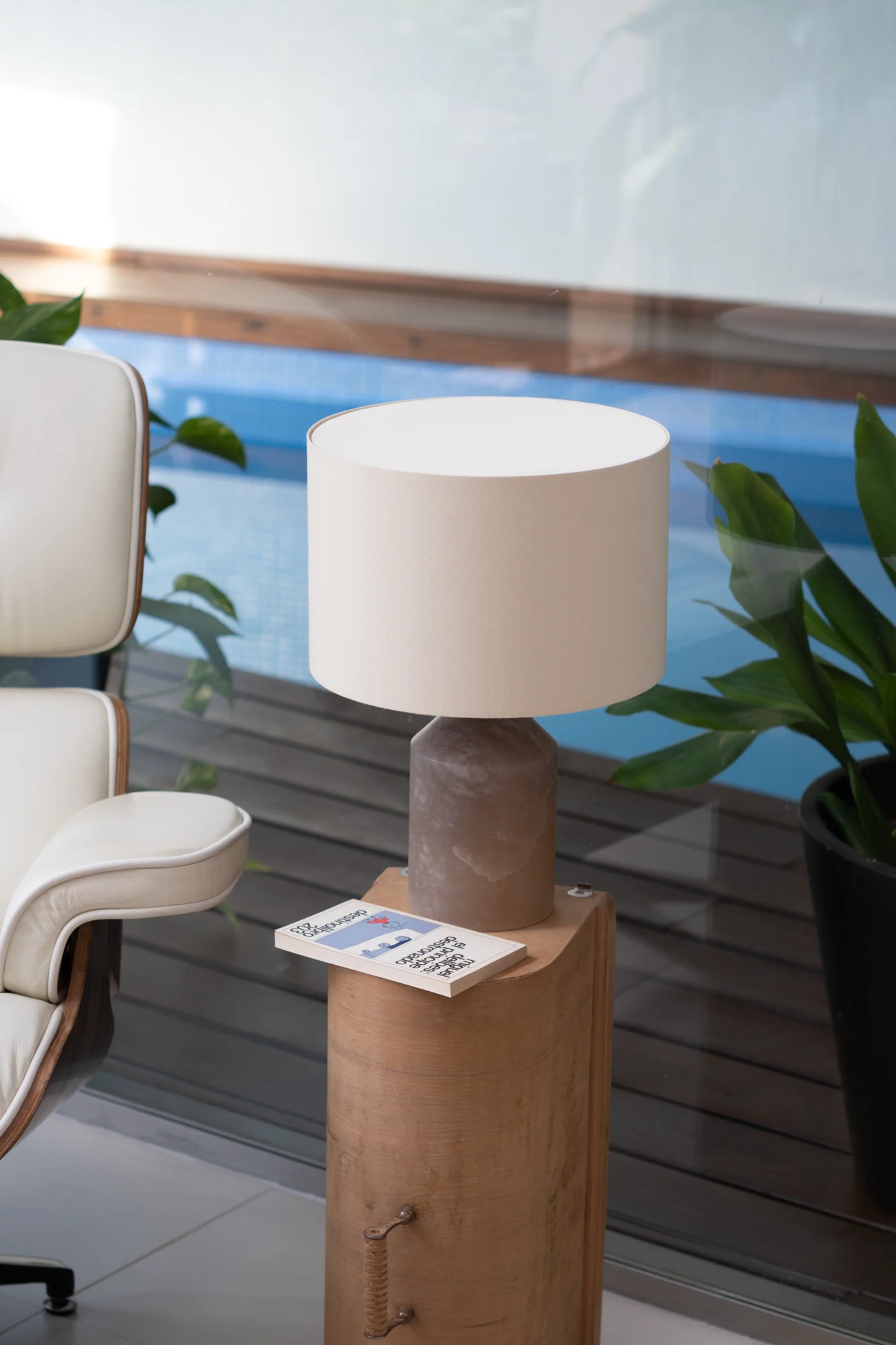 Table lamp with a beige shade on a wooden base next to a chair and plants by a pool.