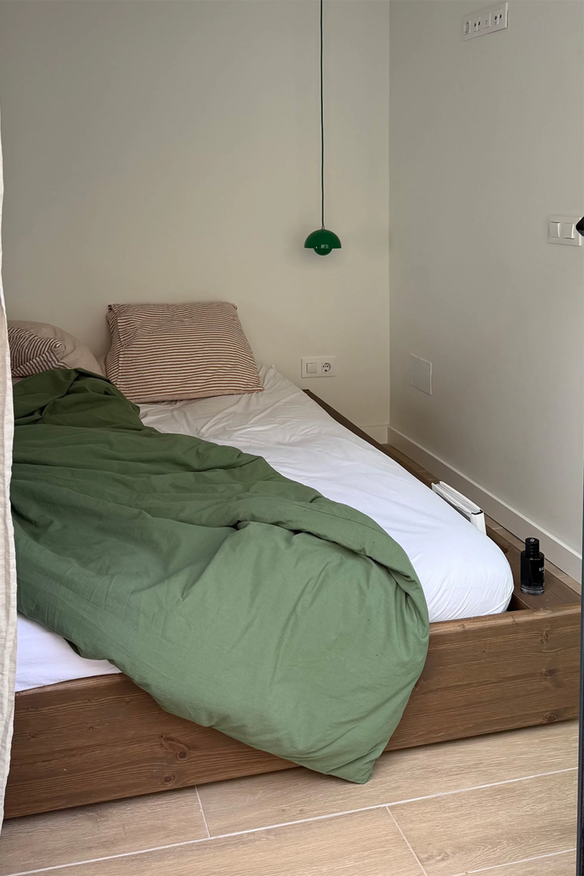 Bed with green blanket and white sheets in a room with beige walls and wooden floor.