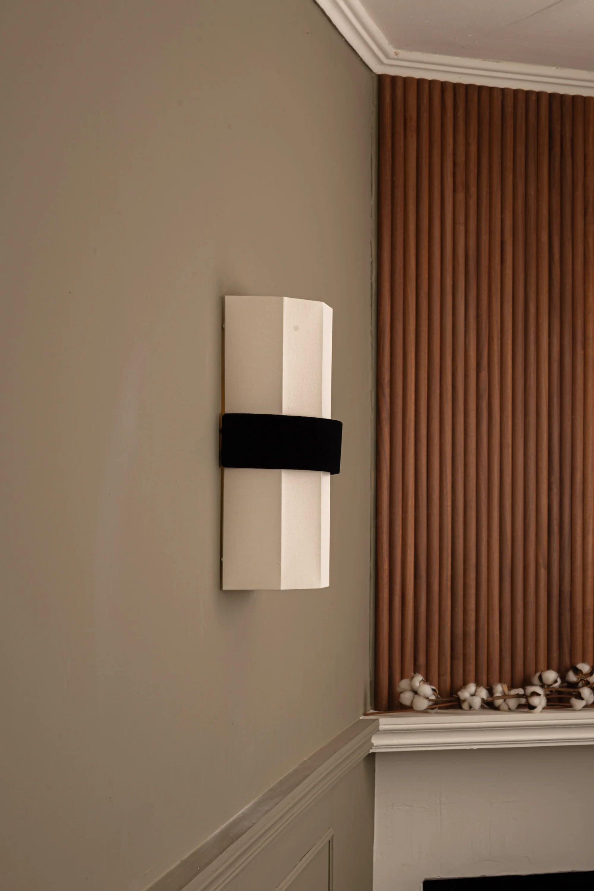 White wall-mounted light fixture with black band on a beige wall next to a window with brown curtains.