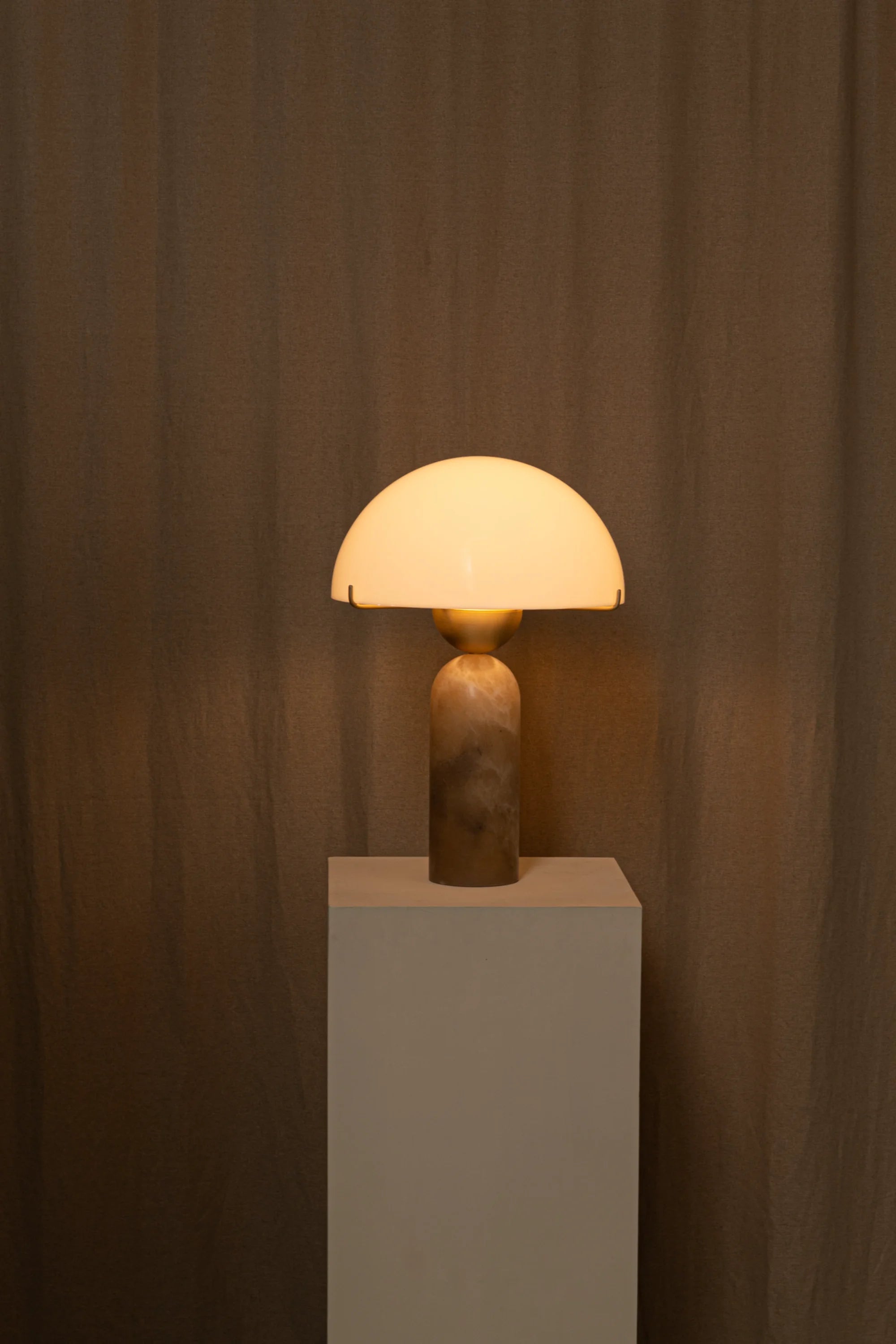 Table lamp with a warm glow on a white pedestal against a brown curtain background