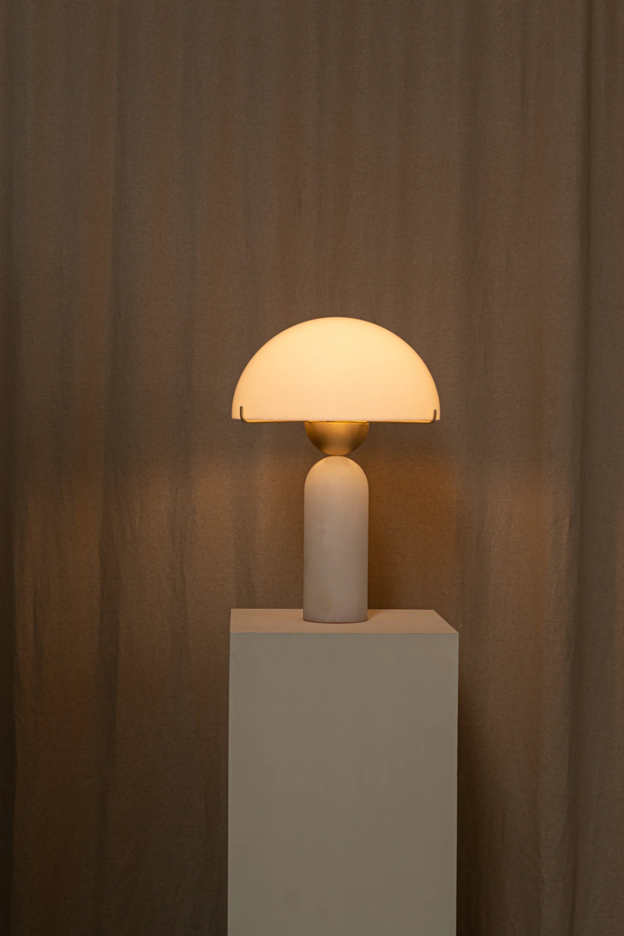 Table lamp with a beige shade on a white pedestal against a brown curtain background
