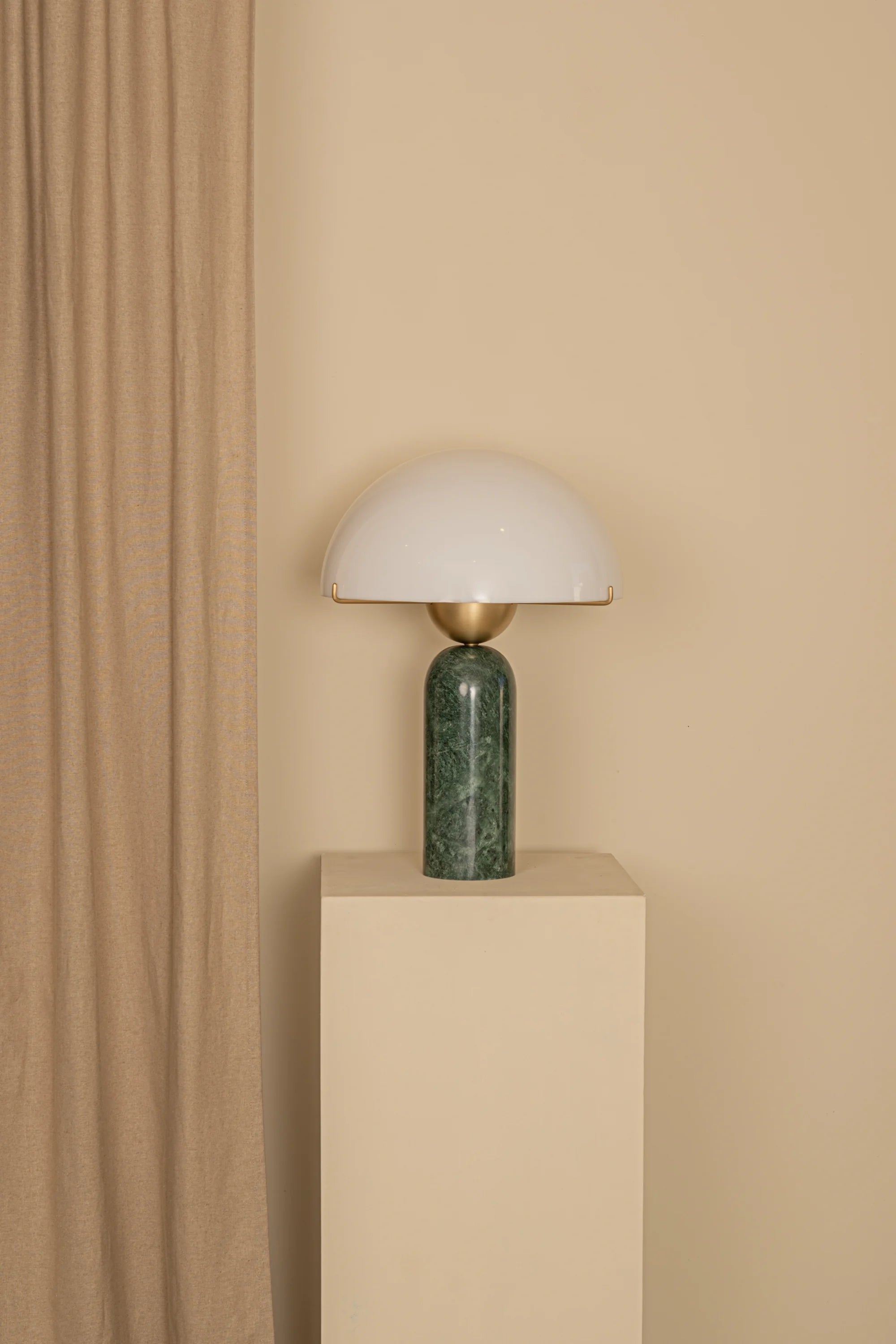 Table lamp with green base and white shade on a beige pedestal against a beige wall.