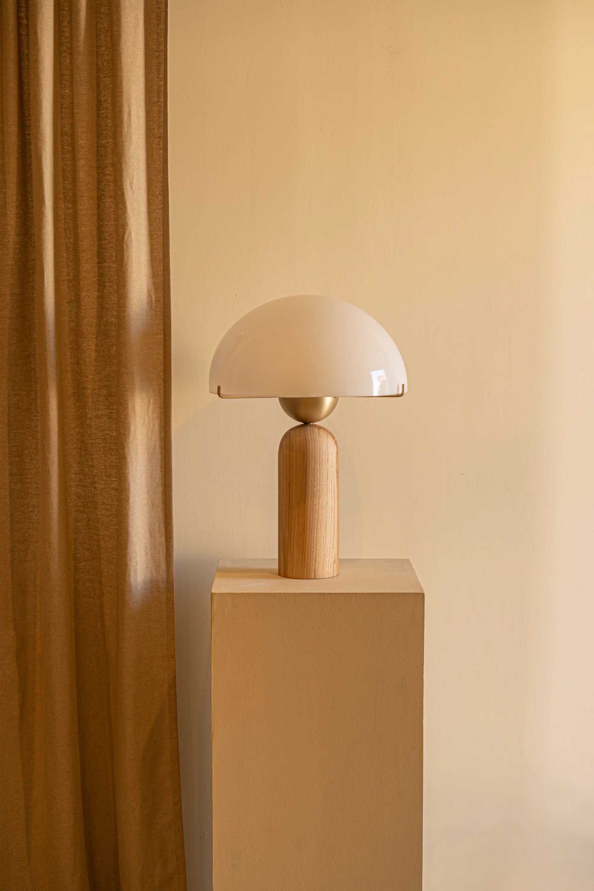 Table lamp with a wooden base and white glass shade on a beige pedestal against a beige wall.