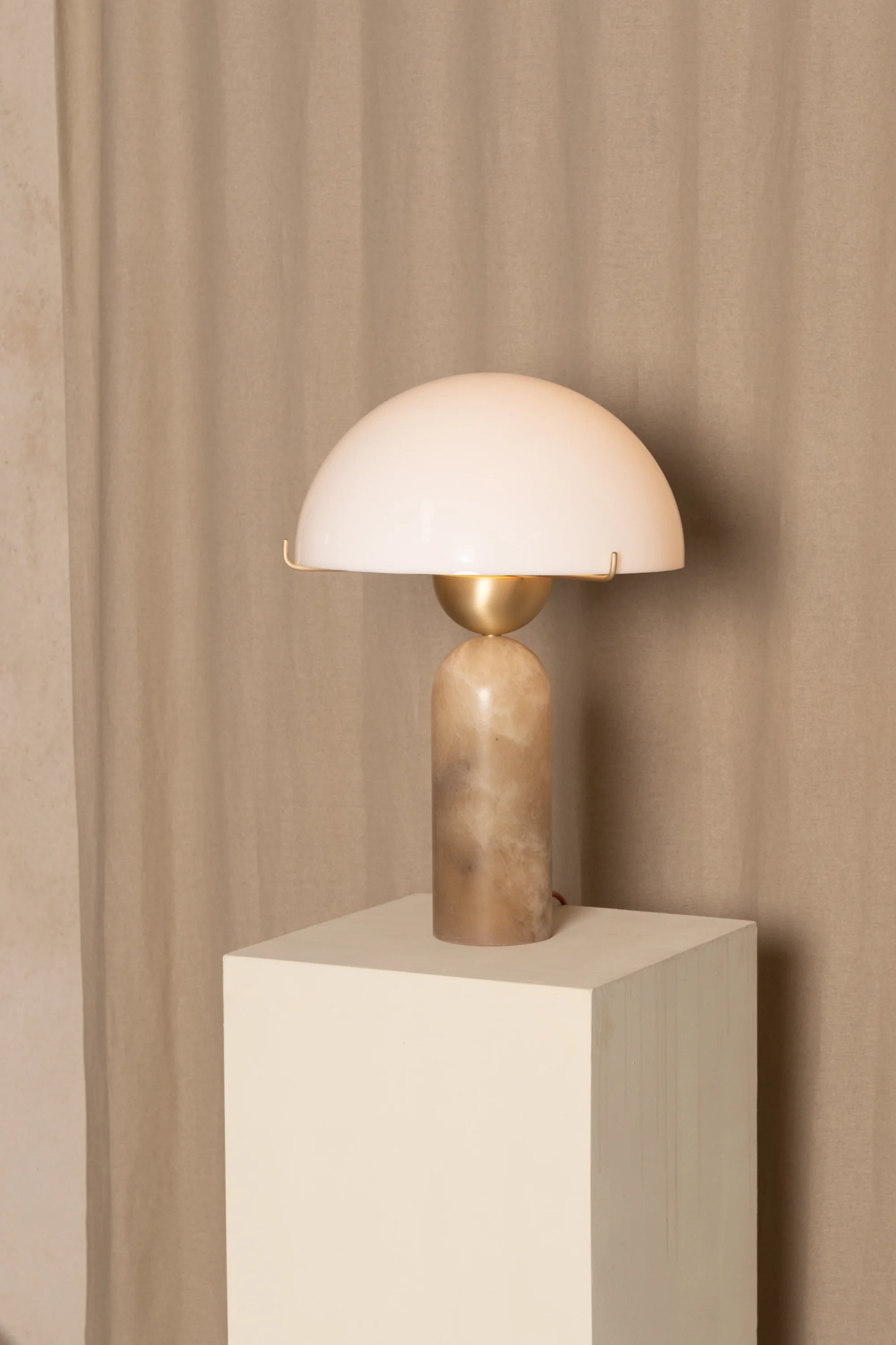 Table lamp with a white shade and bronze base on a beige pedestal against a beige curtain.