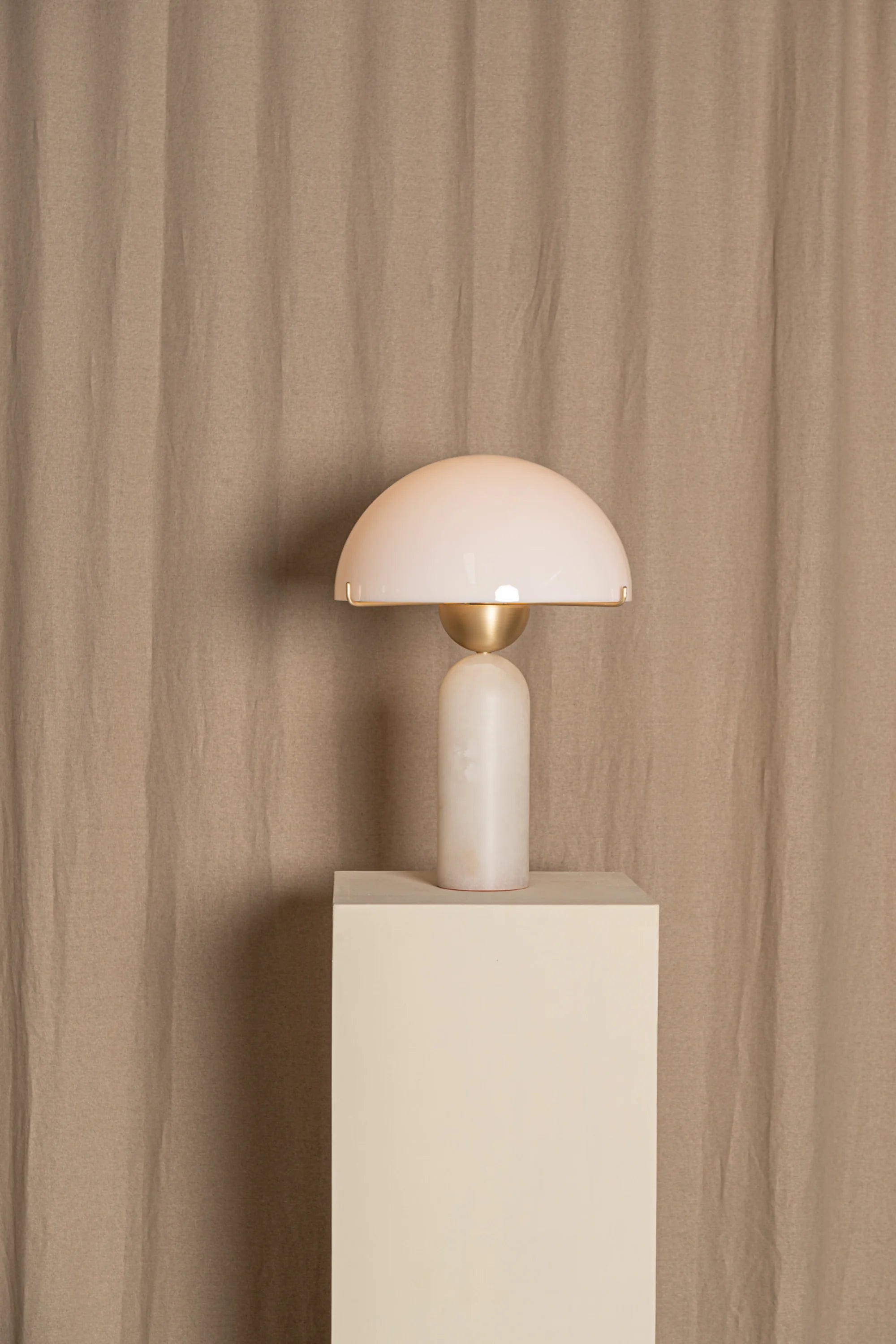 Minimalist table lamp with a white shade and gold base on a beige pedestal against a beige curtain.
