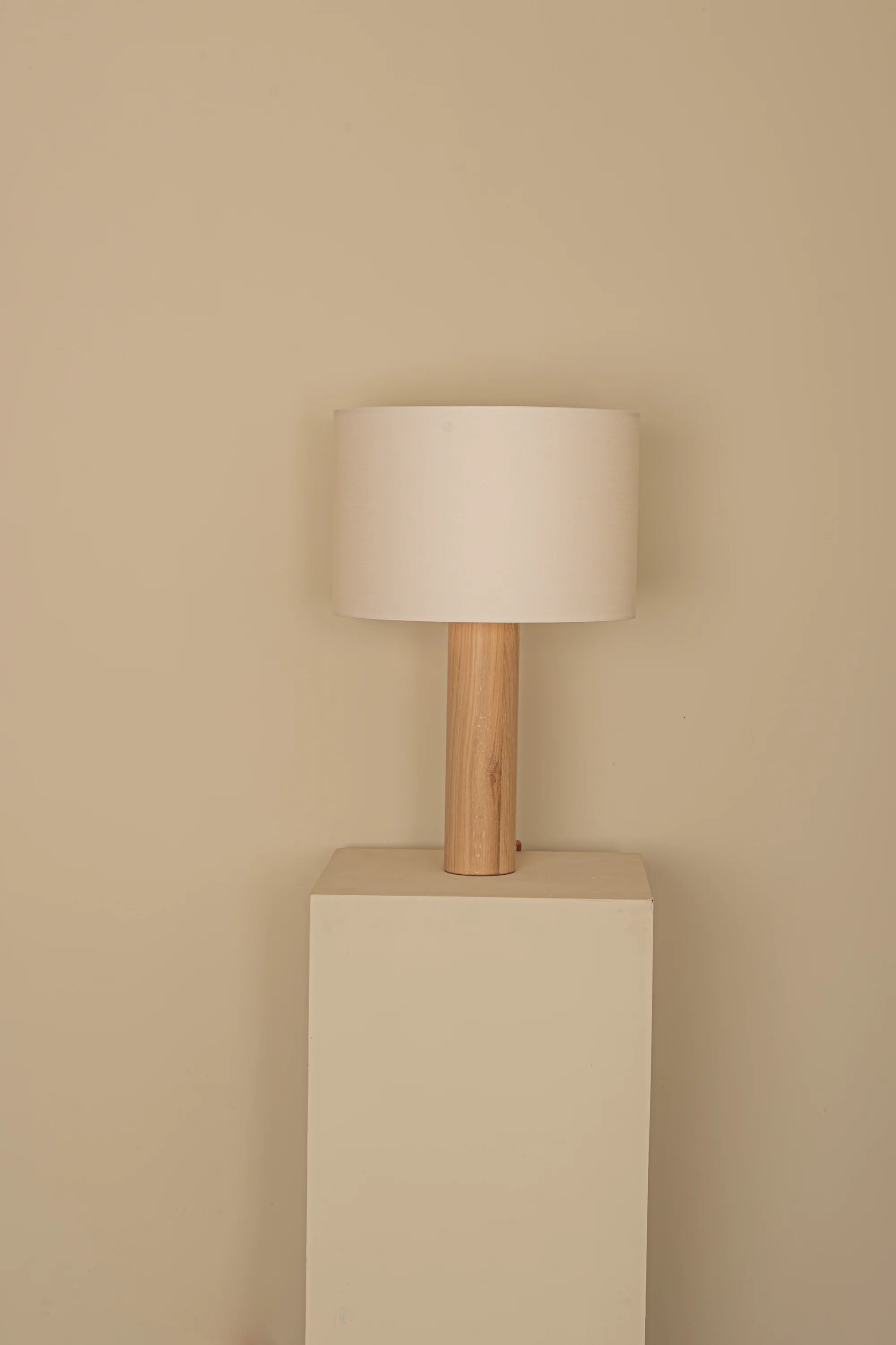 Table lamp with wooden base and white shade on a beige pedestal against a beige wall.