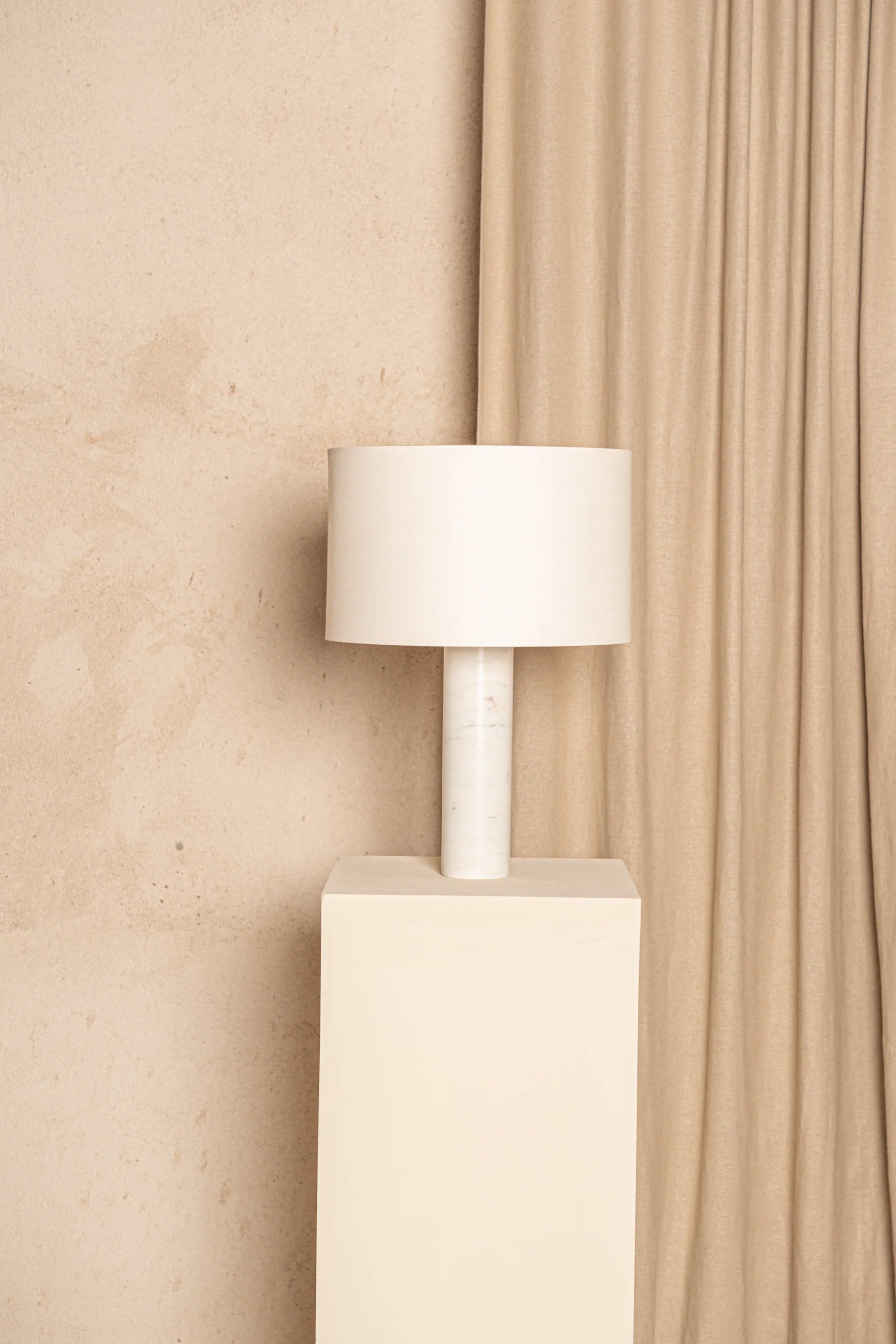 White lamp on a beige pedestal against a beige curtain and wall