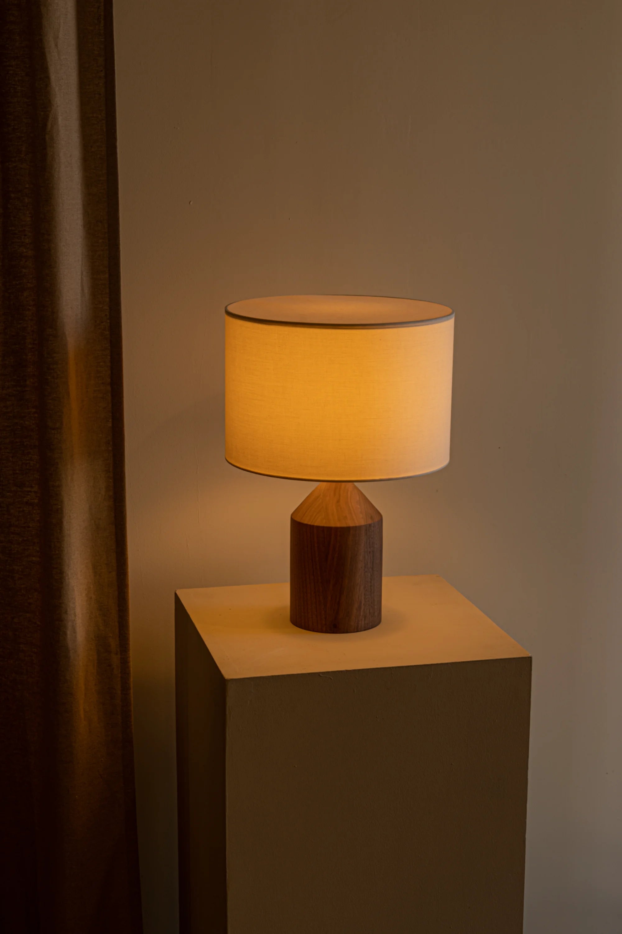 Table lamp with wooden base and beige shade on a square pedestal against a neutral wall.