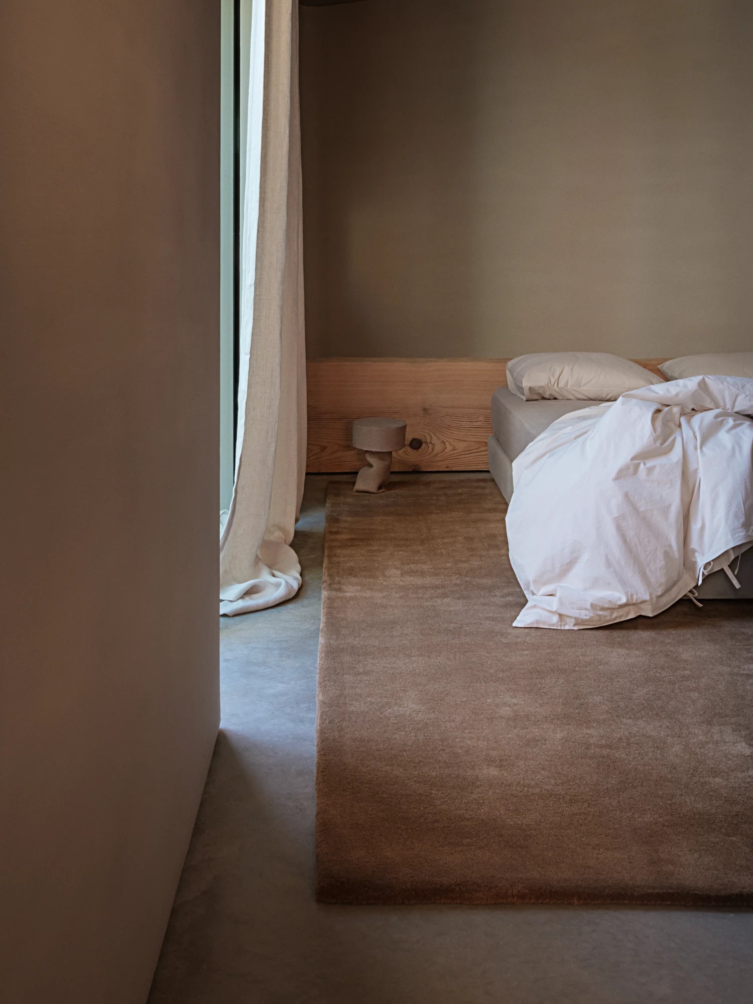 A cozy bedroom features a large, plush, hand-tufted rug in a neutral brown color. The rug is placed under a bed with a wooden headboard and a white comforter. A small, light-colored side table with a lamp is on the rug. The walls are a warm, earthy tone, and a long beige curtain hangs next to a window. The floor is a smooth concrete.