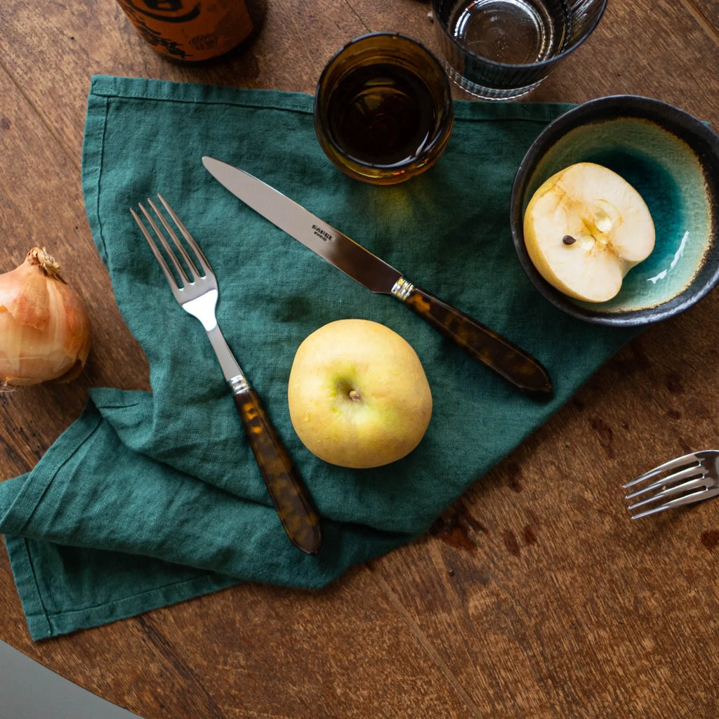Sabre Paris Tortoise Cutlery Set, featuring highly polished stainless steel knife and fork with elegant mottled brown tortoise shell handles, accented by silver rings on a deep teal linen napkin. This stylish cutlery set is ideal for adding a touch of vintage charm to special occasion dining or everyday meals.