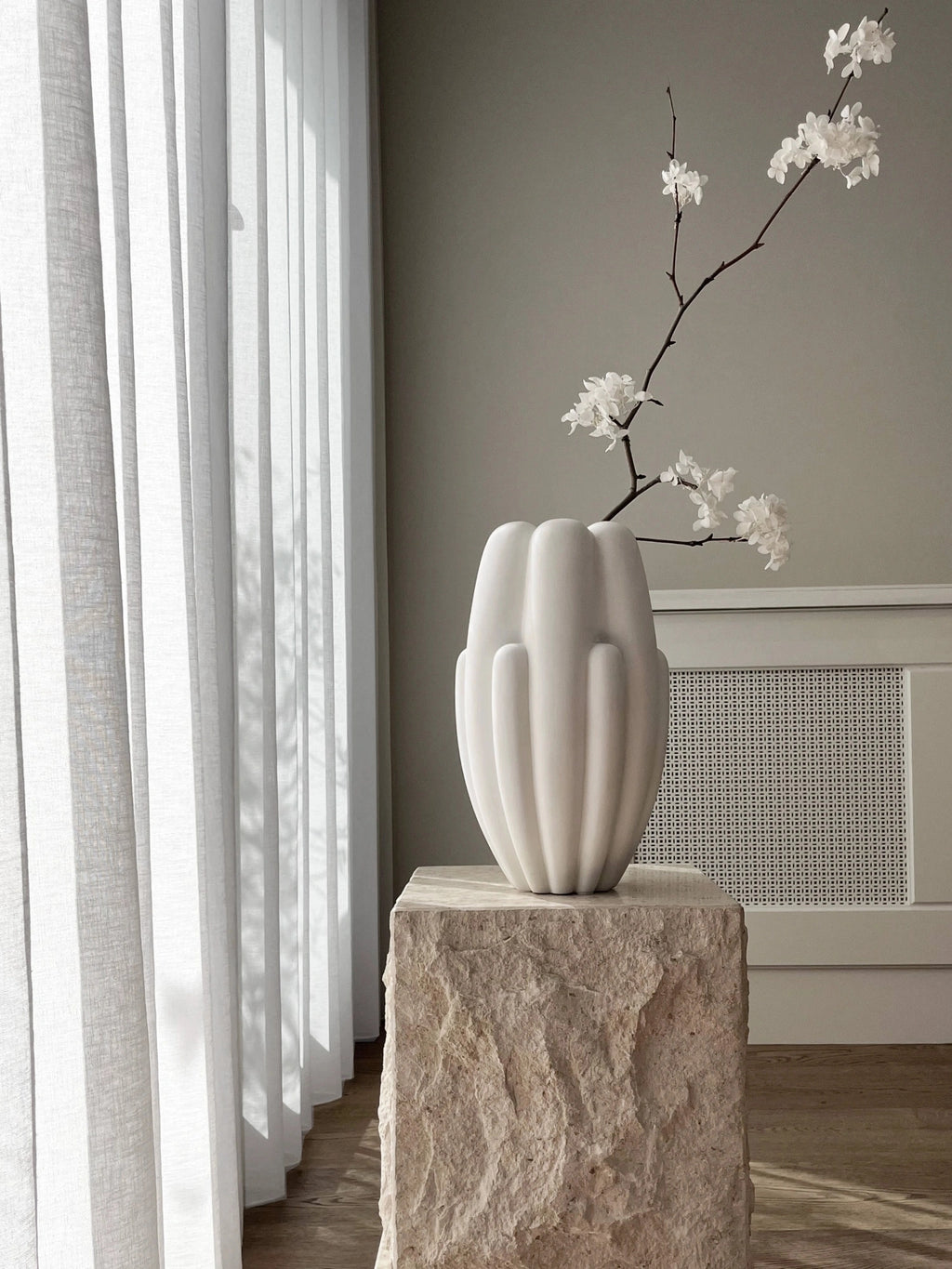 101 CPH Bloom Slim Vase in a modern, matte off-white ceramic, featuring a ribbed, organic form. Displayed on a rough-hewn stone pedestal with white blossoming branches, it's a premium decorative accent. Ideal for contemporary interior styling.