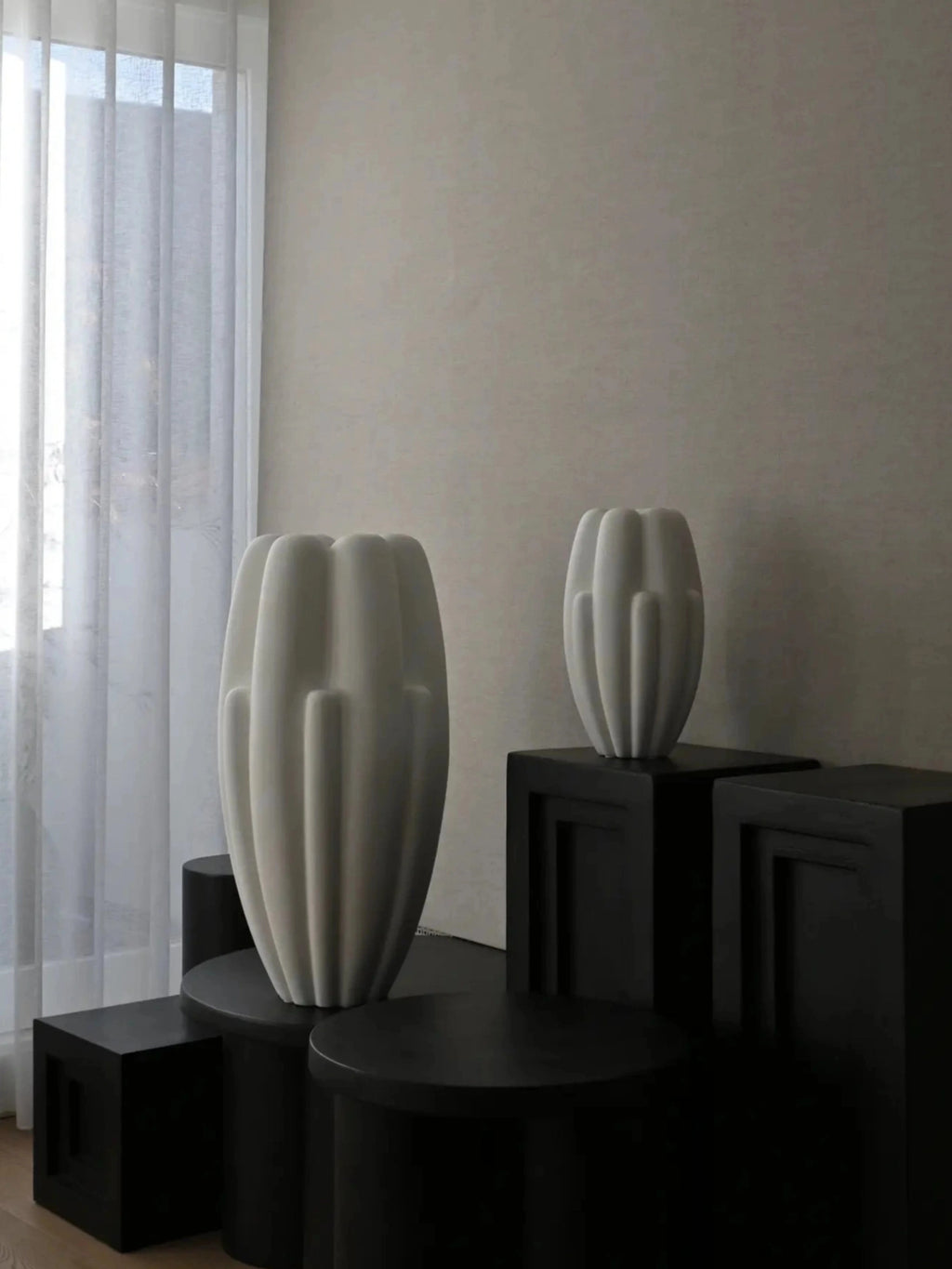 101 CPH Bloom Slim Vase. Two off-white ceramic vases with distinctive ribbed, organic shapes displayed on black sculptural pedestals against a light wall and sheer curtain. A premium Scandinavian design piece.