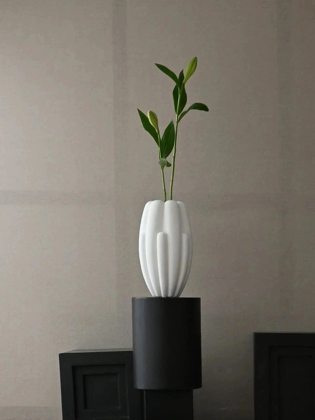 101 CPH Bloom Slim Vase in white ceramic, featuring a ribbed, organic form. This premium decorative vase holds green foliage, displayed on a black stand. Perfect for a modern, minimalist interior.