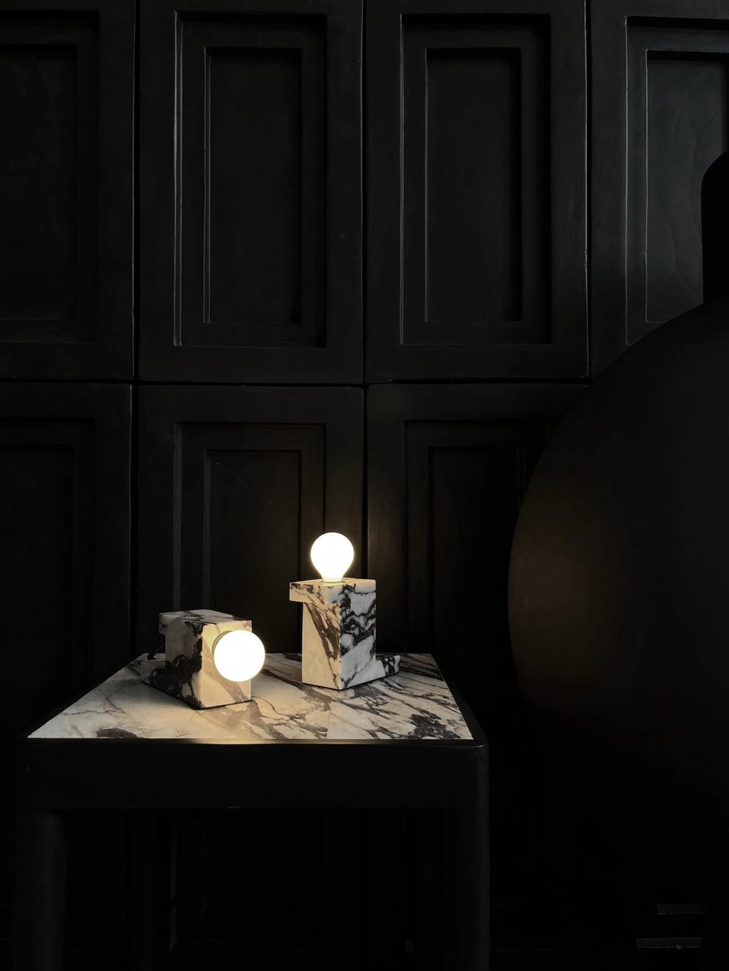 101 CPH Brick Lamp: Two luxury black and white marble lamps with round white bulbs, displayed brightly lit on a matching marble table against a dark paneled wall. Perfect for modern interior lighting.