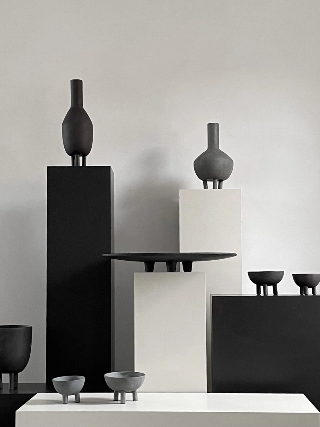 101 CPH's Duck Tray in dark grey ceramic, with an oval shape and three distinct feet, showcased among other minimalist vessels on white and black pedestals. Ideal for modern Scandinavian decor.