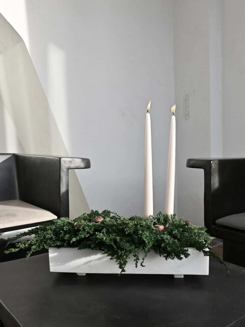 101 CPH Formalism Tray on a black table, presenting an arrangement of evergreen foliage, pinecones, and two lit tall, slender cream candles in a minimalist, rectangular white concrete tray. This authentic, handcrafted piece creates a stylish center.