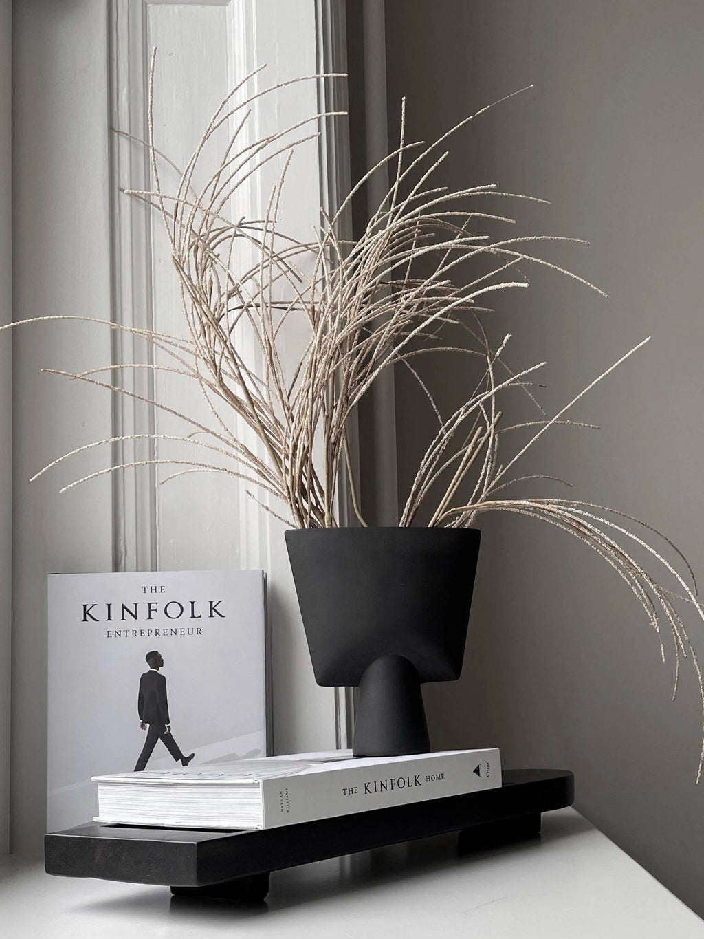 101 CPH Geta Tray, a minimalist black rectangular wood serving tray with two subtle feet, styled with two Kinfolk books and a modern black vase holding dried white grass. Its luxury design makes it perfect for refined home decor.