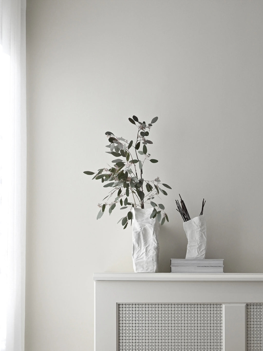 101 CPH Kami Vase, a set of two unique handcrafted crumpled white stoneware vases, displayed with greenery and branches on a white mantelpiece for a chic, modern aesthetic.