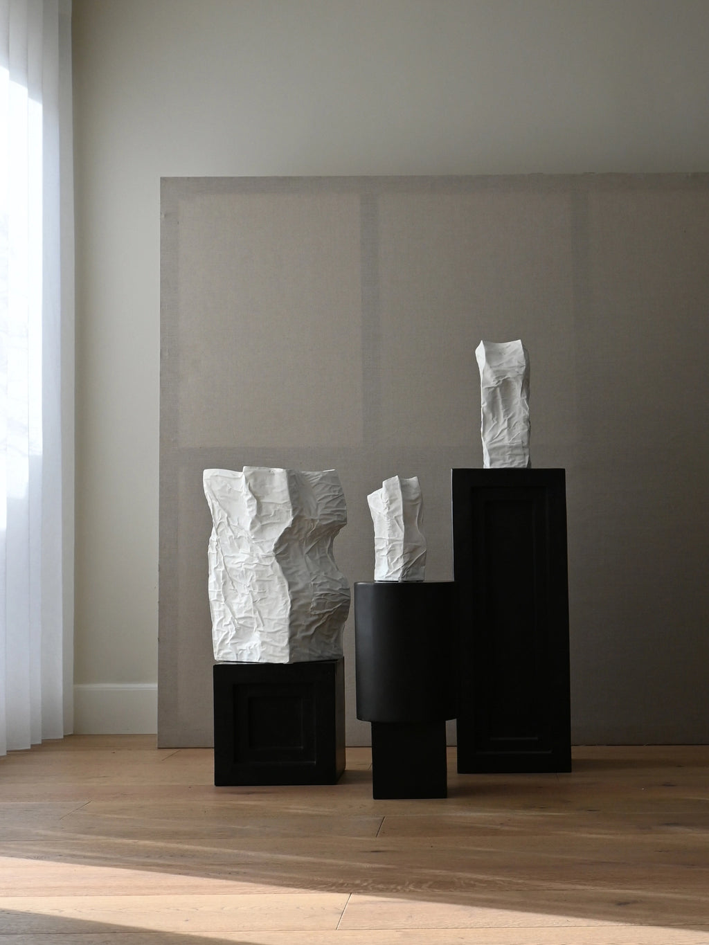 101 CPH Kami Vase, a set of three unique crinkled white ceramic vases with organic, paper-like textures, displayed on black pedestals in a minimalist room. Ideal for creating a modern focal point.
