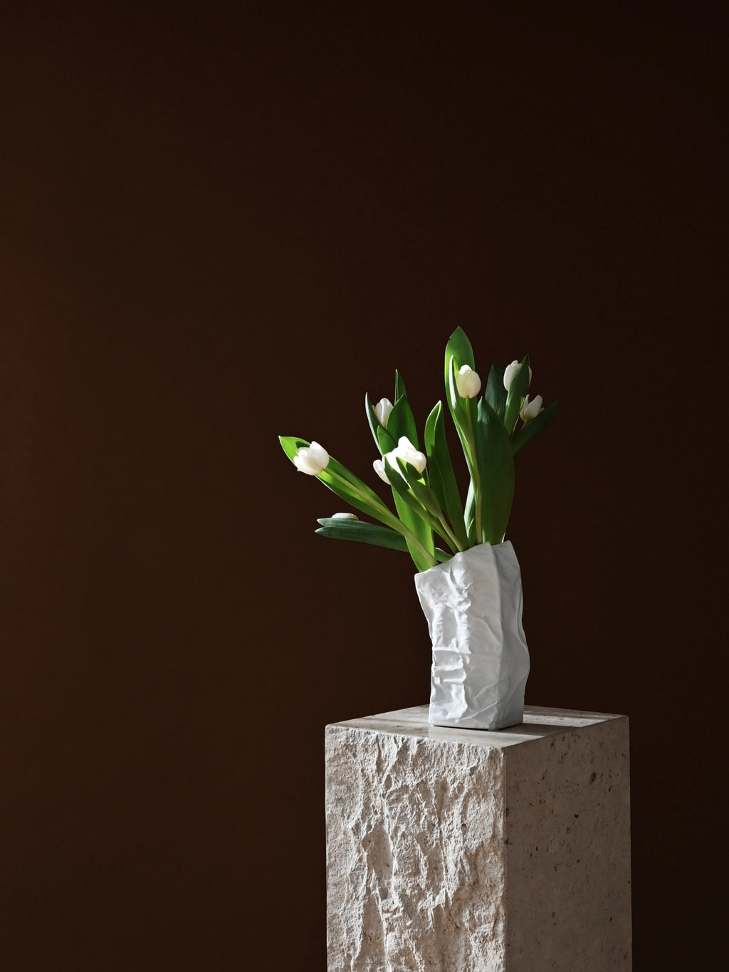 101 CPH Kami Vase, a premium white ceramic vase with an organic, crumpled paper bag texture, stands on a rough grey stone pedestal with white tulips. Ideal for modern minimalist decor.