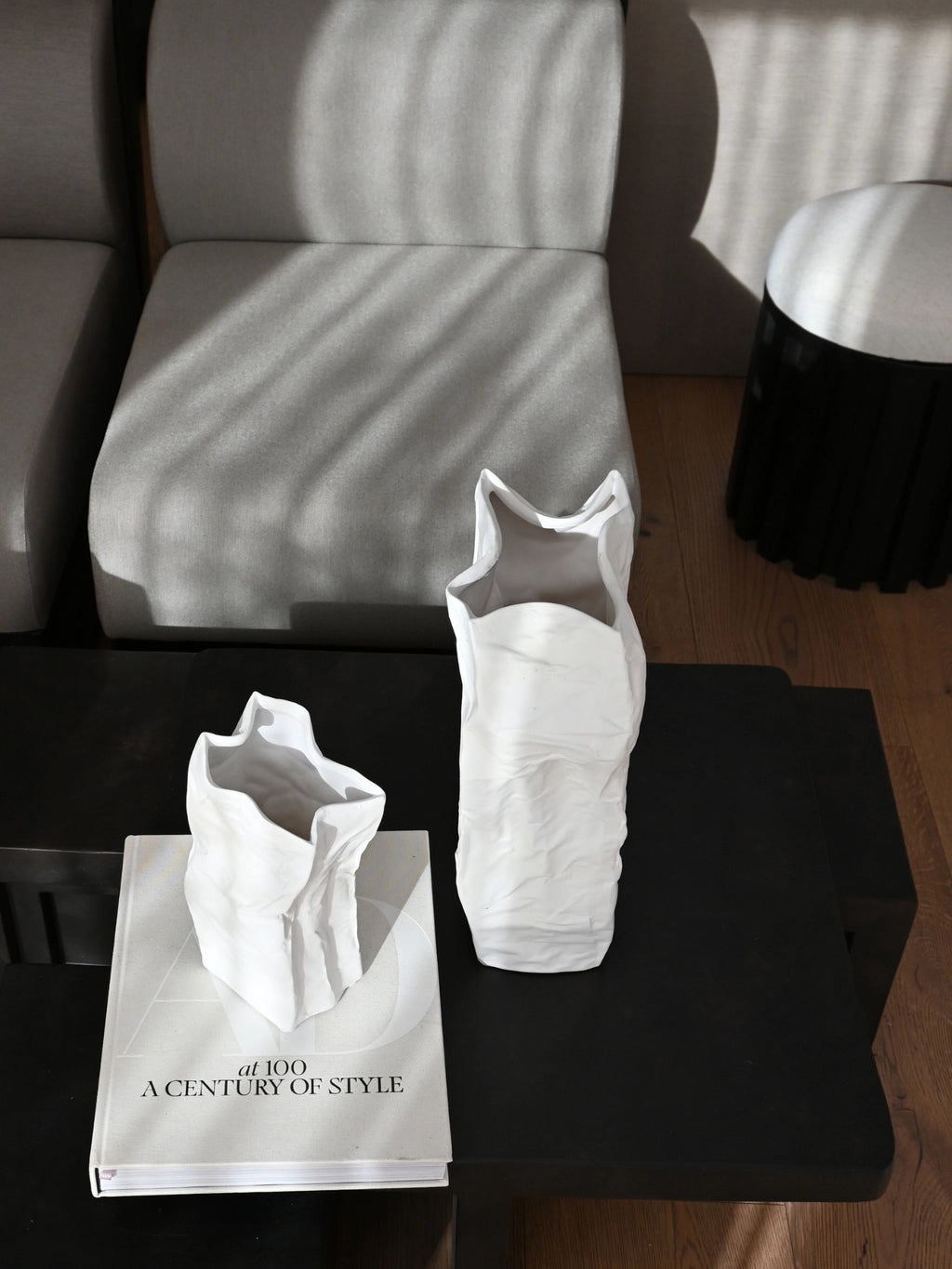 101 CPH Kami Vase. Two premium white textured ceramic vases resembling crumpled paper, styled on a dark table with a light grey couch in a modern interior. Ideal for contemporary home decor.
