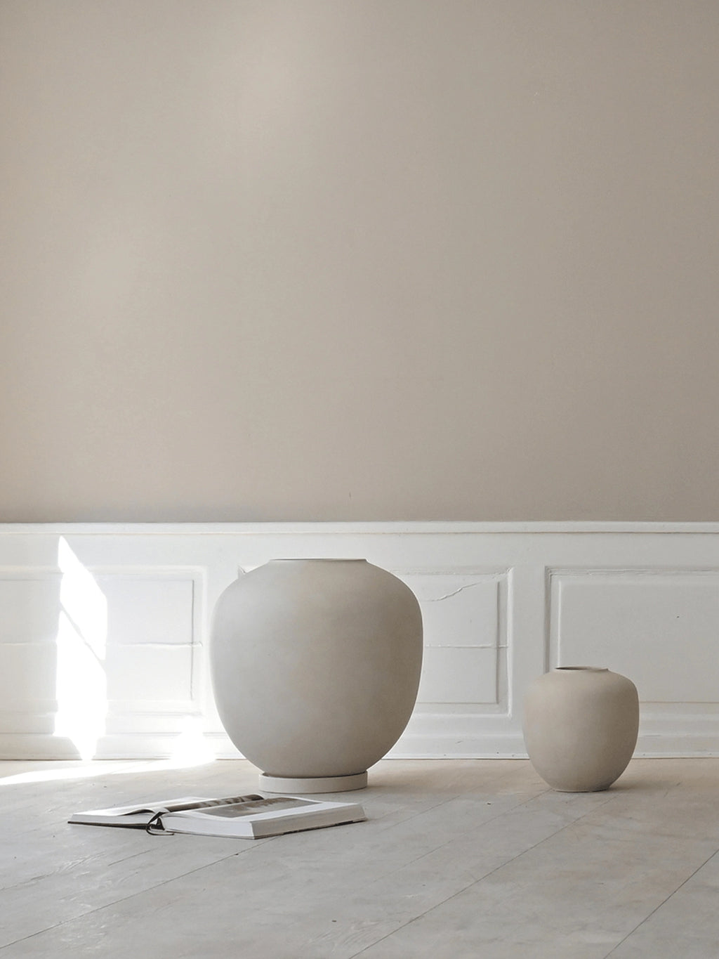 101 CPH Sunao Planter, two modern matte beige ceramic planters of varying sizes with subtle texture, displayed on a pale wood floor with an open book. A luxury home decor accent.