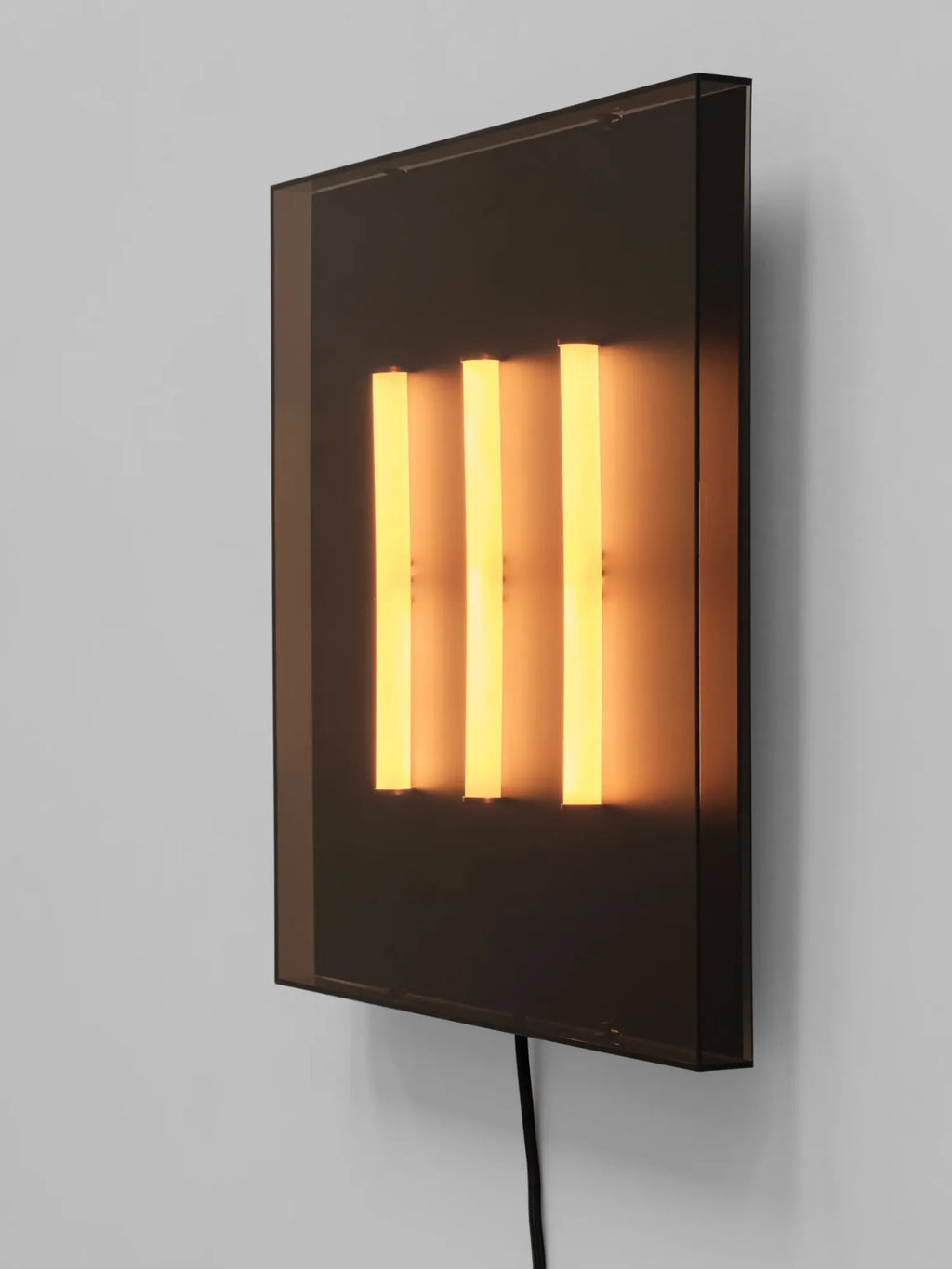 101 CPH Trois Wall Lamp, a sleek rectangular dark smoked glass panel mounted on a light gray wall, with three vertical warm amber glow light bars illuminating through. An authentic designer wall light.