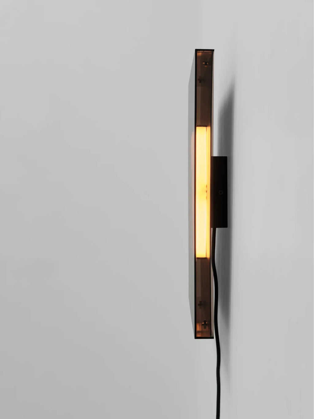 101 CPH Trois Wall Lamp: Rectangular, dark smoked glass and black painted steel luxury wall light with a warm yellow glow, mounted on a light gray wall. A modern design statement.