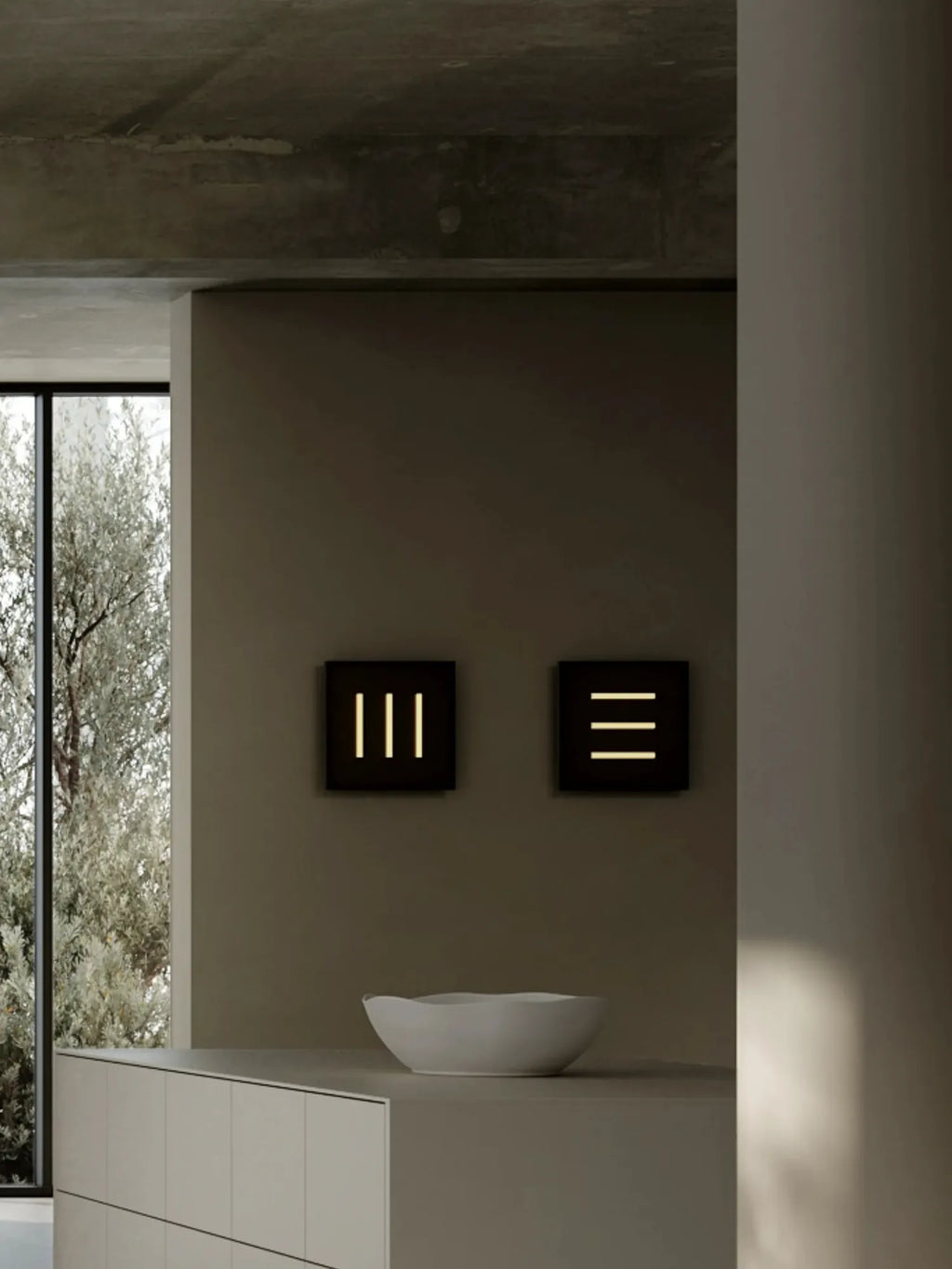 101 CPH Trois Wall Lamp: Two modern black square wall sconces are mounted on a dark grey wall, each emitting warm yellow light through three vertical or horizontal slits, styled above a light grey kitchen island with a white bowl. A premium designer light for contemporary interiors.