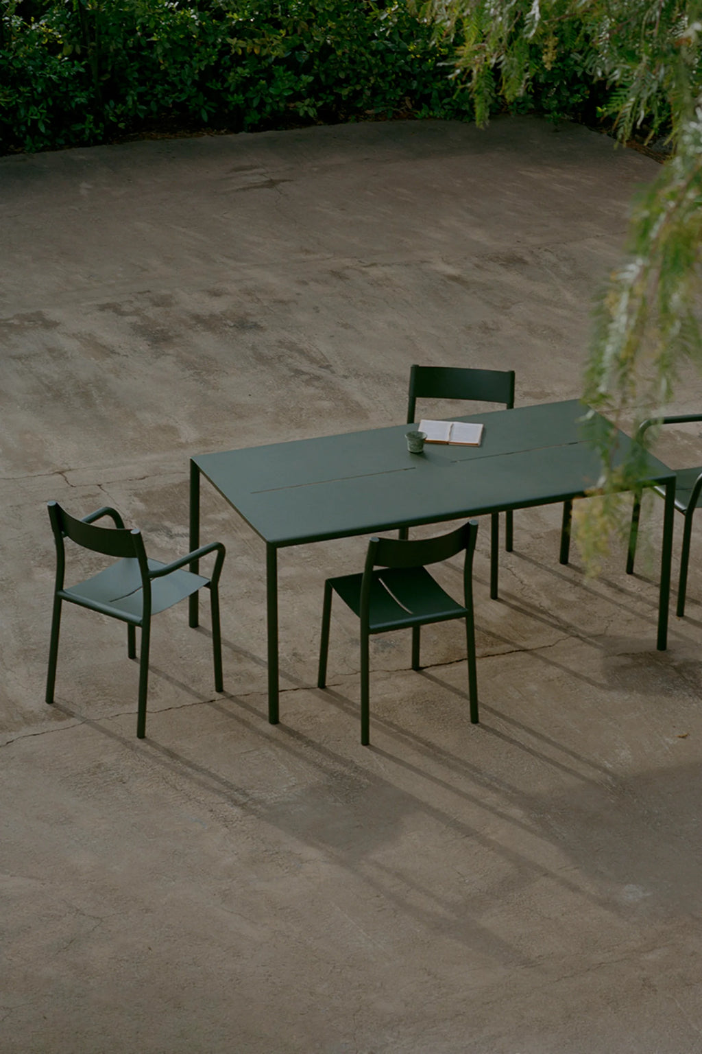 New Works Outdoor May Furniture Set for 4 Persons, a sleek modern minimalist outdoor dining set crafted from durable matte dark green powder-coated metal. The rectangular dining table pairs with four matching chairs, featuring a clean contemporary design with slender frames and a distinctive slot in the seat. Ideal for a sophisticated Scandinavian-inspired patio, garden dining area, or modern deck furniture.