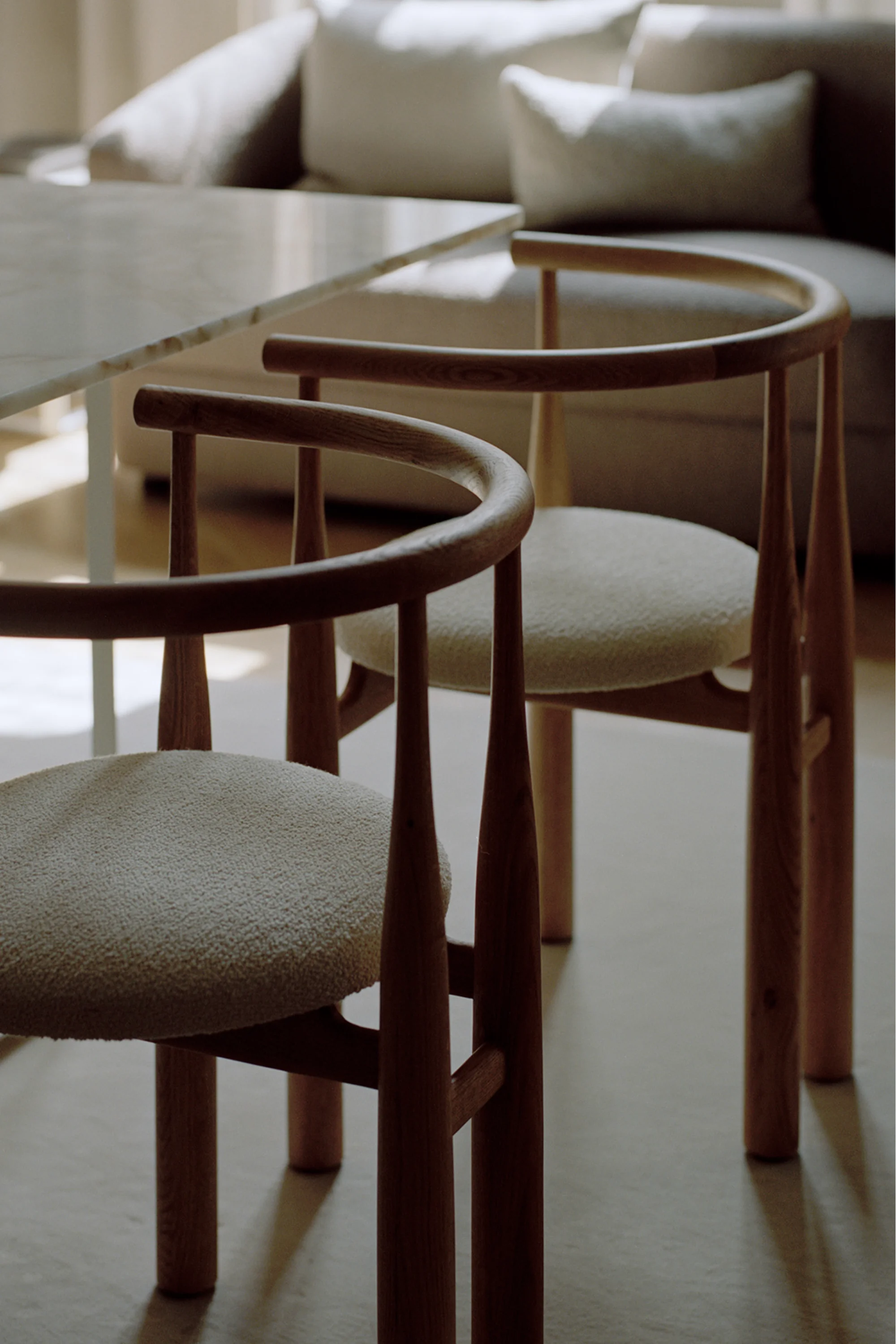 New Works Bukowski Dining Chair: Two natural oak chairs with distinct curved backrests and light cream bouclé upholstered seats, positioned in a contemporary dining setting. Ideal for sophisticated interiors.