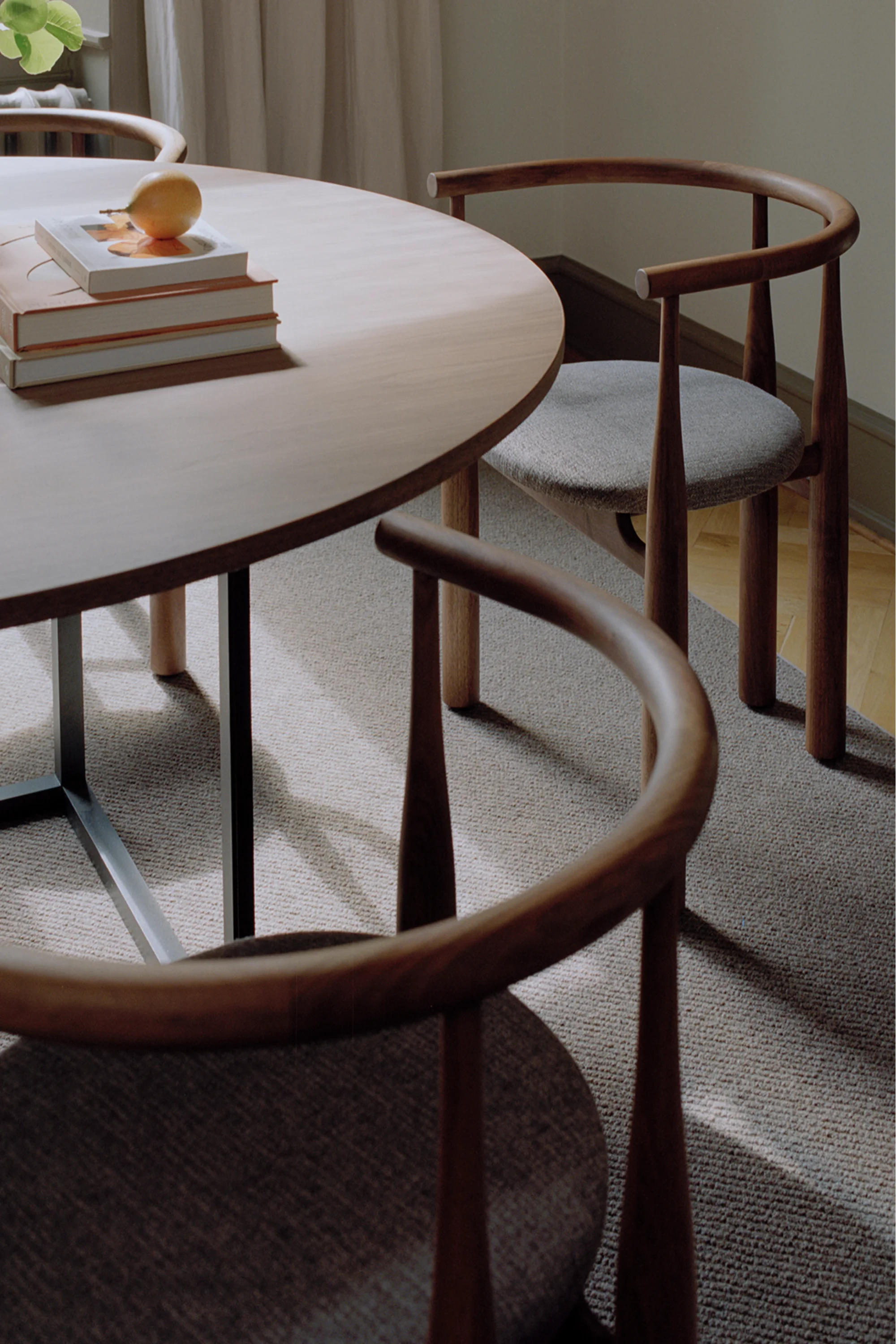 New Works Bukowski Dining Chair in a natural walnut wood finish with round, curved backrest and light grey upholstered seat, displayed next to a matching light brown round dining table. A premium, comfortable seating solution.