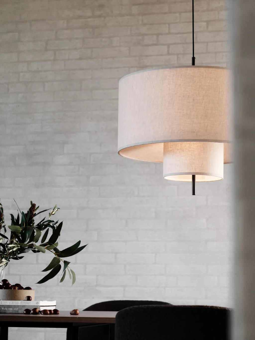 New Works Margin Pendant Lamp, shown hanging in a lifestyle shot. This premium tiered lamp features two cream-colored fabric shades and a central black stem for a soft, diffused glow. Ideal for modern dining or living spaces.