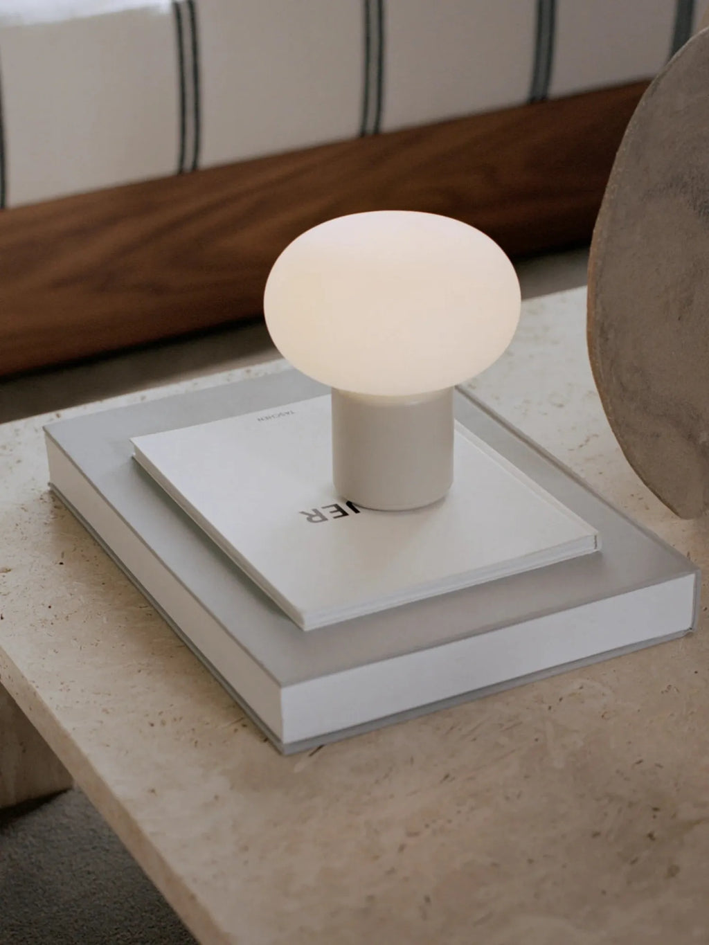 New Works Karl-Johan Portable Table Lamp: A modern mushroom-shaped lamp with an opaque white glass top and a matte white cylindrical base, styled on two books atop a light brown marble surface. This premium wireless lamp is ideal for versatile ambient lighting.