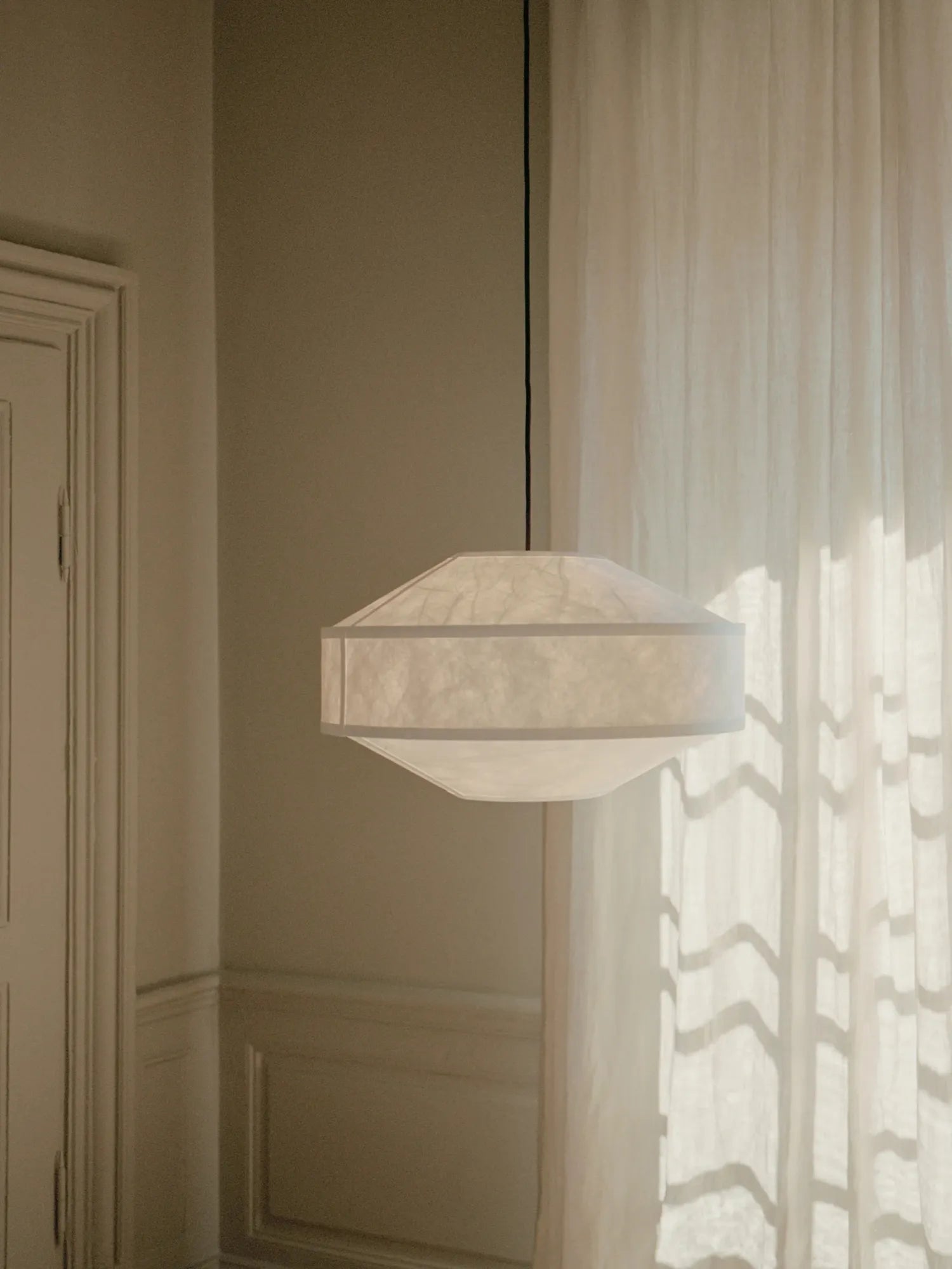 New Works Kite Pendant Lamp: A minimalist white fabric shade with an angular, textured surface, suspended from a black cord in a soft, sunny room. Ideal for modern ambient lighting.