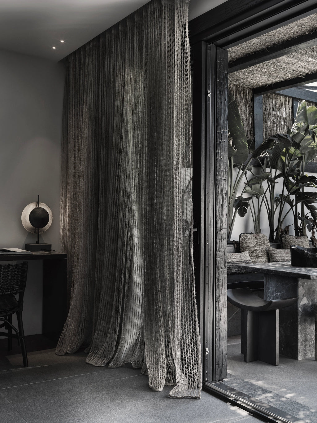 101 CPH Dusk Table Lamp, a luxury black metal sculptural lamp with a glowing round amber light, displayed on a dark wooden desk in a modern interior setting. Offers moody atmospheric lighting.