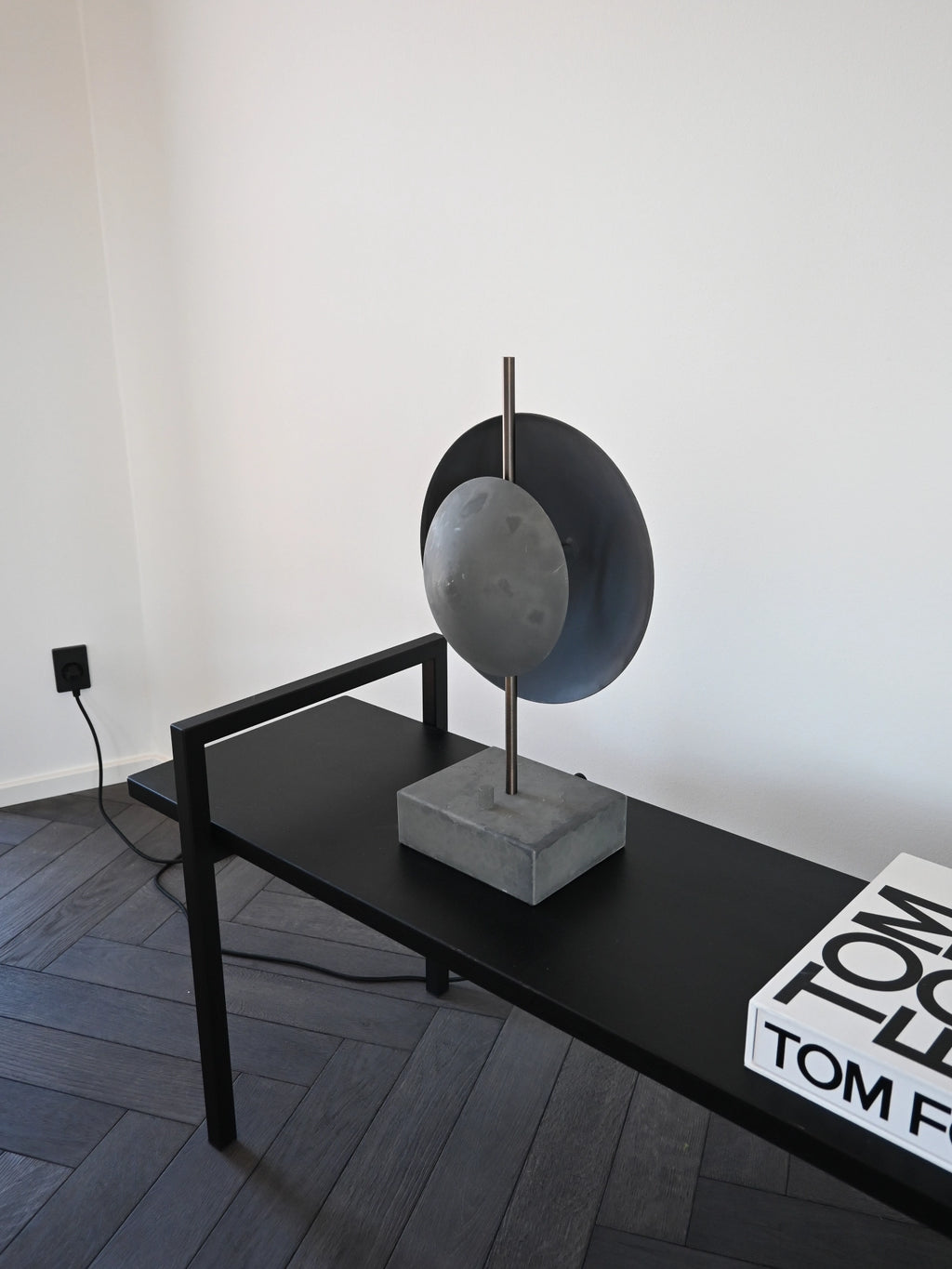 101 CPH Dusk Table Lamp, a contemporary modern design featuring a dark grey concrete base, an antique brass stem, and two overlapping matte dark grey metal discs, styled on a black console table. Ideal for adding a designer accent.