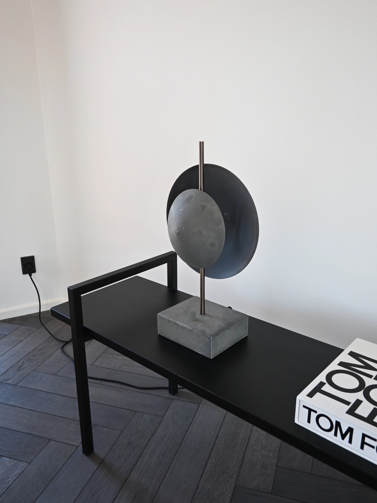 101 CPH Dusk Table Lamp, a contemporary modern design featuring a dark grey concrete base, an antique brass stem, and two overlapping matte dark grey metal discs, styled on a black console table. Ideal for adding a designer accent.