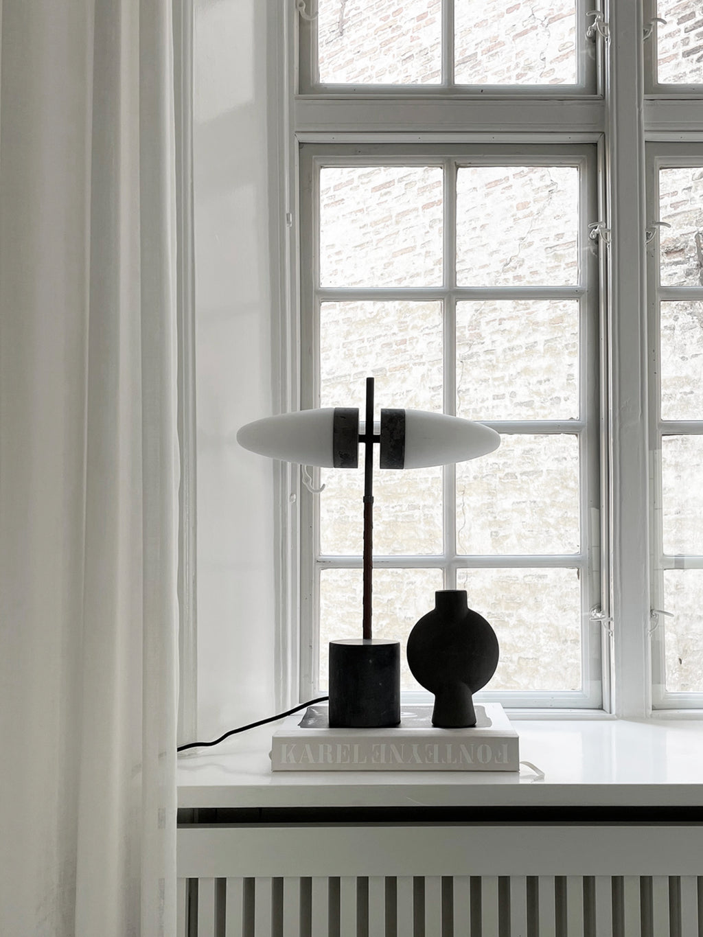 101 CPH Bull Table Lamp features an elongated matte white glass diffuser with dark bronze accents, set against a window with white curtains. A luxury statement piece for any modern interior.
