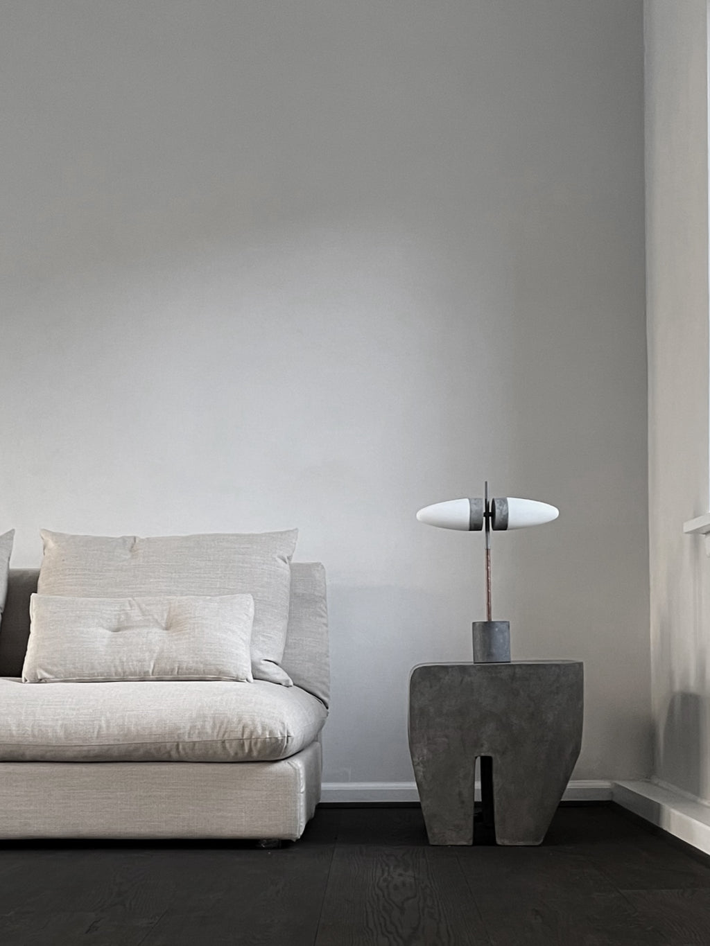101 CPH Bull Table Lamp, a luxury concrete and bronze sculpture-like lamp with two white oval shades, styled on a dark grey concrete side table beside a light grey sofa. Perfect for modern living spaces.