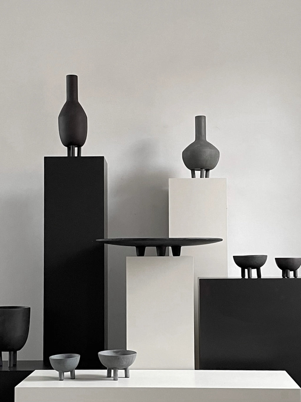 101 CPH Duck Bowl collection in varied grayscale studio display. Multiple textured, dark gray ceramic bowls, vases, and dishes, some footed, showcased on matte black and white pedestals. Premium decorative accents for modern interiors.