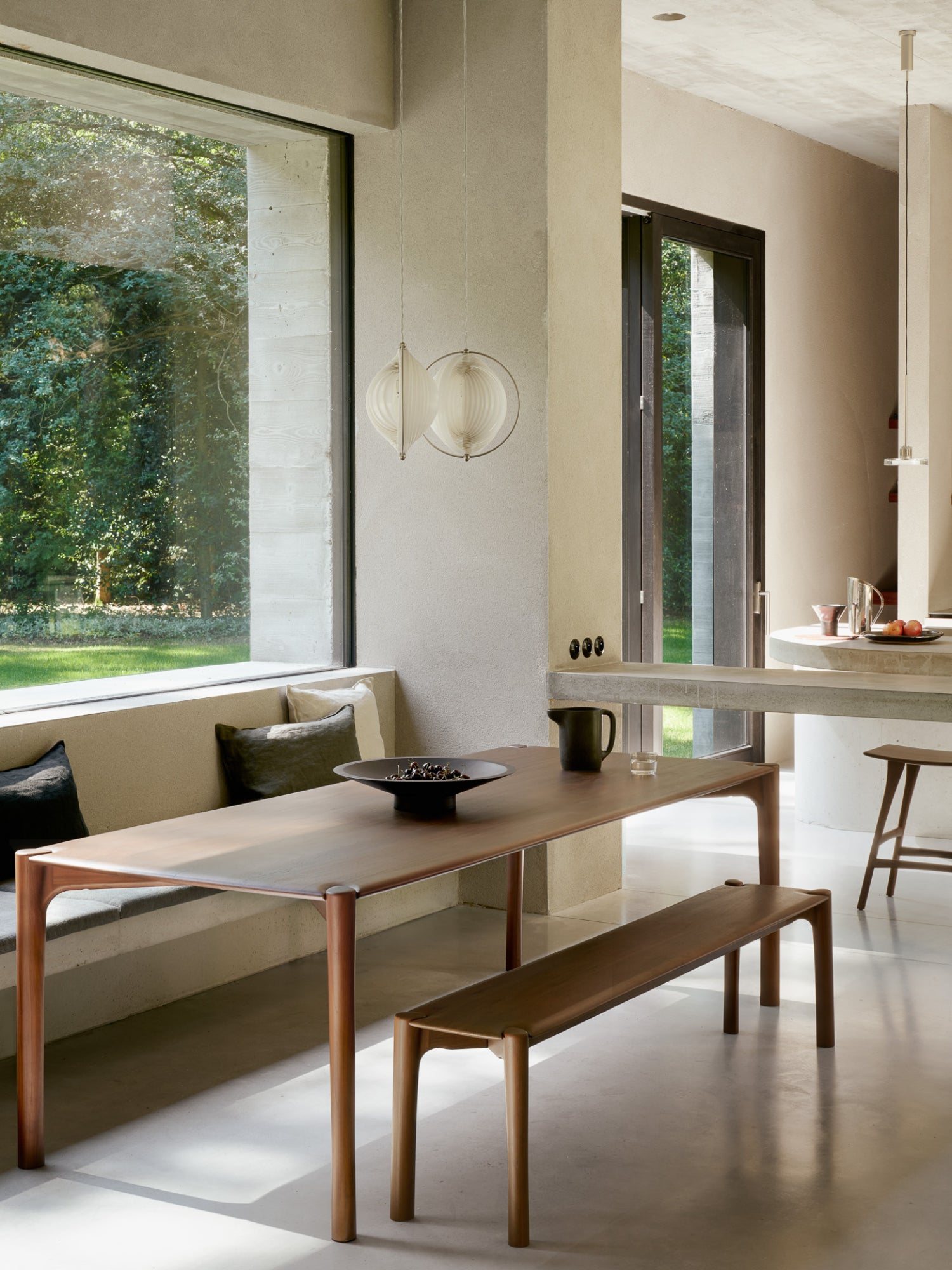 Ethnicraft PI Bench in a modern dining area, crafted from rich brown teak wood, positioned under a window with a matching dining table, creating an elegant, minimalist aesthetic.