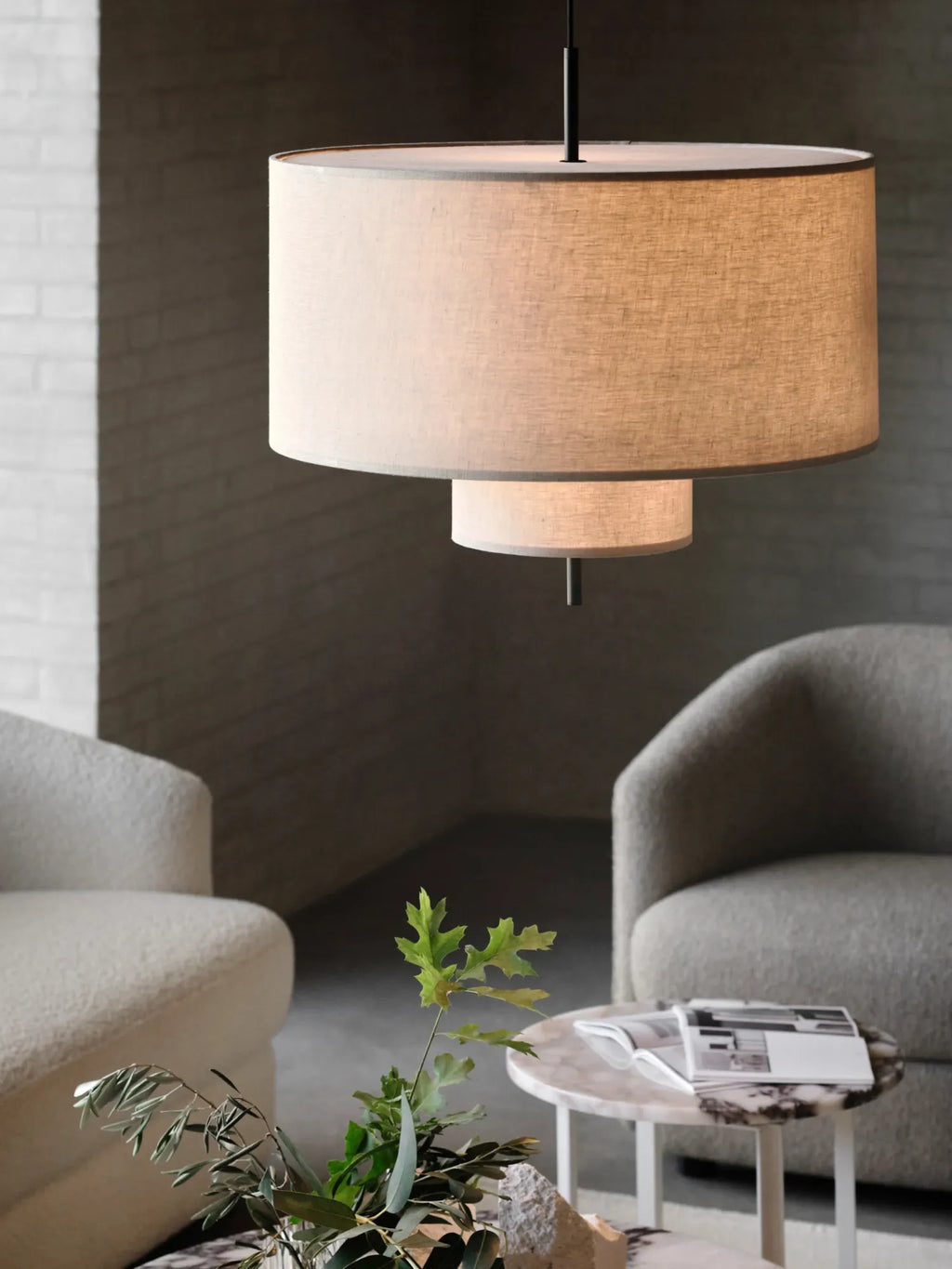 New Works Margin Pendant Lamp, featuring two tiered, natural linen drum shades with slim gray edges, suspended from a black rod in a stylish living room setting. This premium designer lighting casts a warm, ambient glow. Ideal for sophisticated interiors.