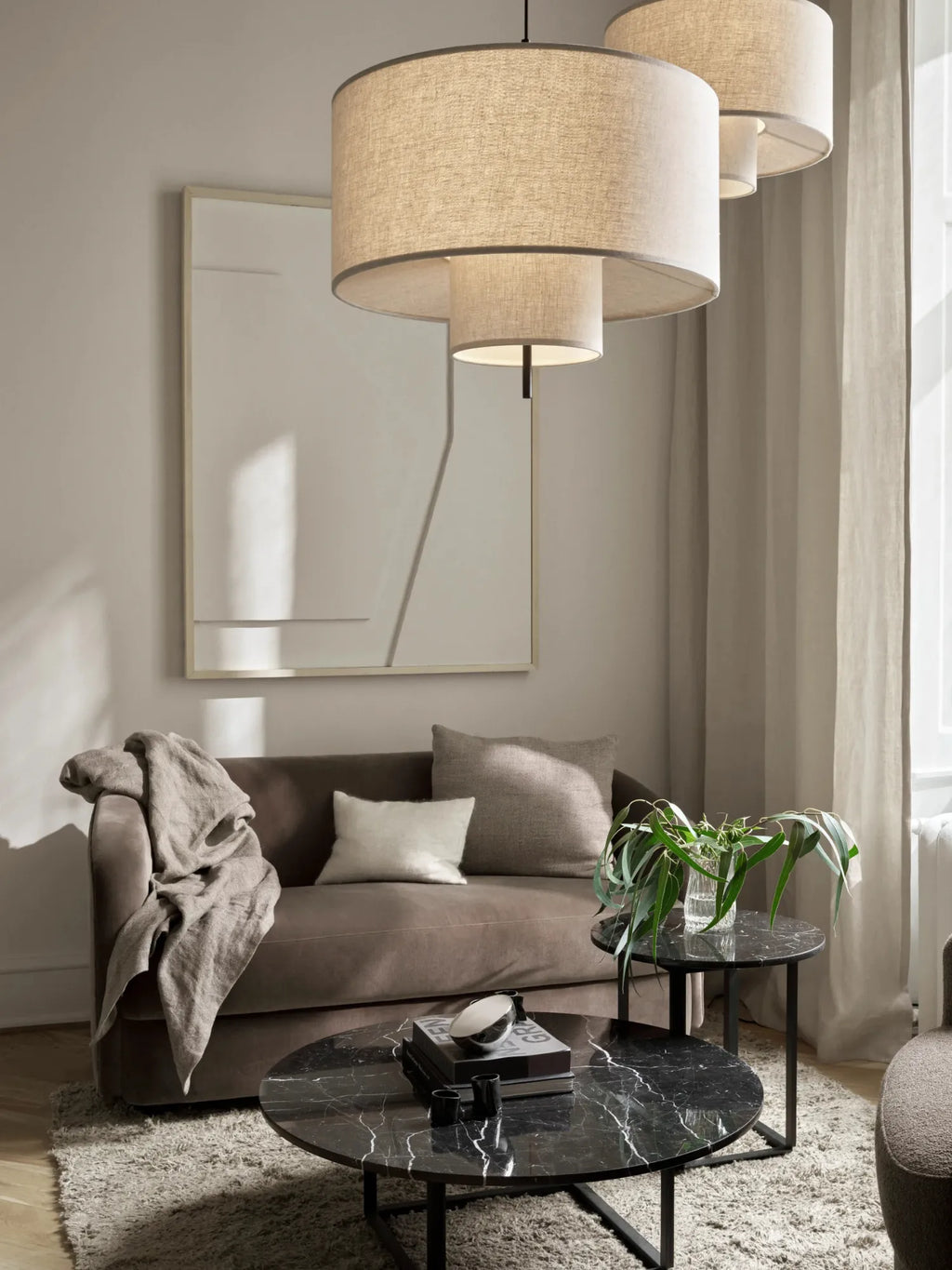 New Works Margin Pendant Lamp, photographed in a modern living room setting, features tiered beige linen shades with dark trim, offering soft, diffused light. This elegant, premium lighting piece enhances any contemporary space. Ideal for refined ambient lighting.