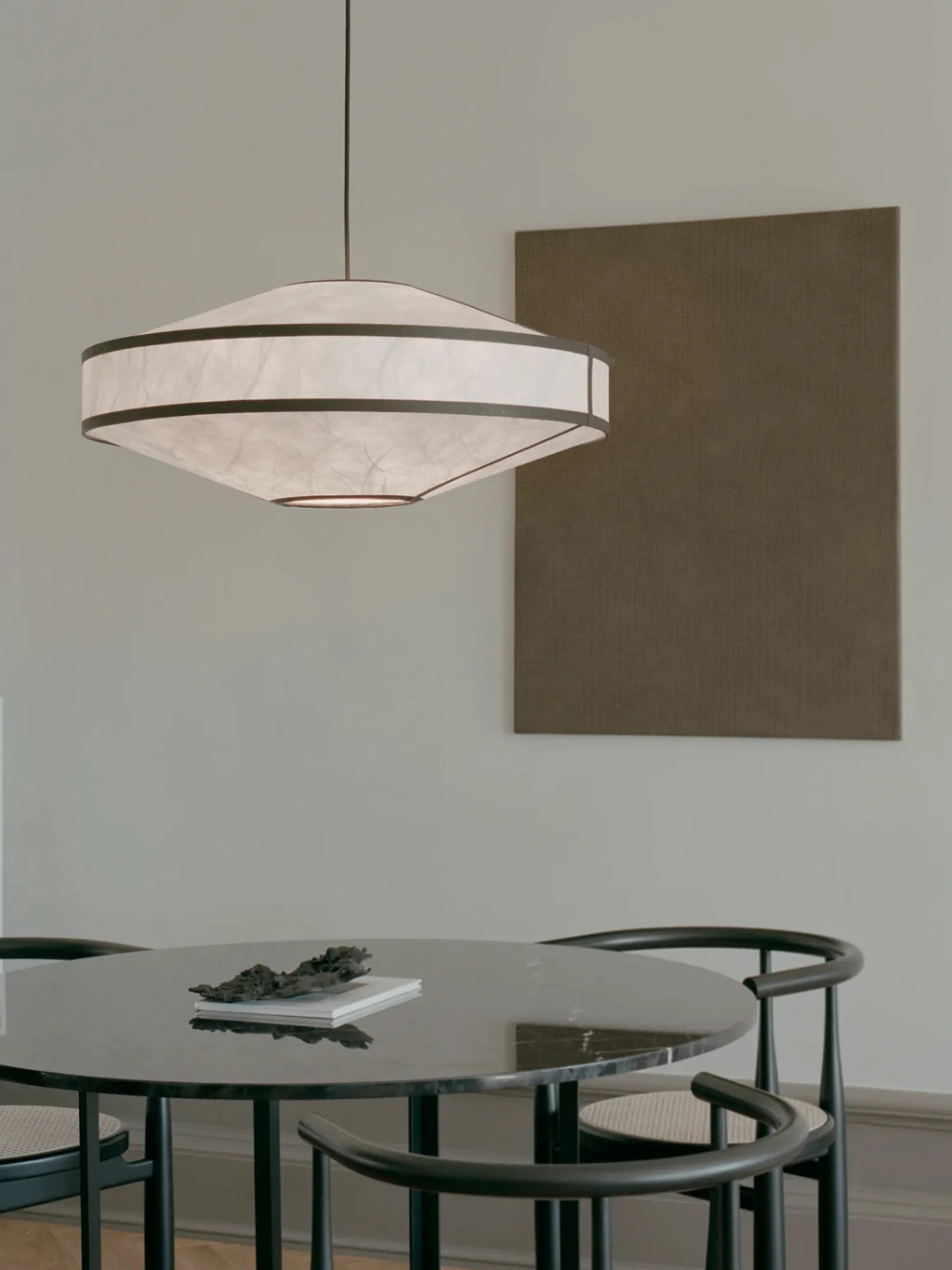 New Works Kite Pendant Lamp; a unique, modern off-white rice paper shade with black trim, hangs centered above a sleek black marble dining table and chairs in a minimalist room. Ideal for contemporary living spaces.