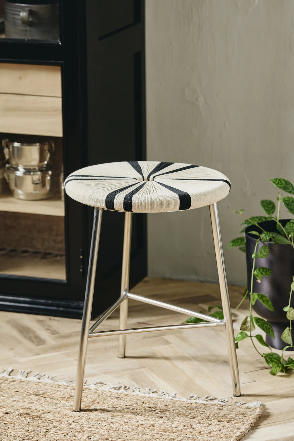 Nordal Morice Stool, a striking modern accent stool featuring a round seat intricately hand-woven with cream-colored rope and bold black radial stripes, creating a captivating graphic pattern. It stands on sleek, polished silver metal legs with a sturdy triangular crossbar, offering a contemporary industrial chic aesthetic. This versatile woven rope stool is perfect as a stylish entryway seat, bedroom accent, or a unique addition to modern living room decor.