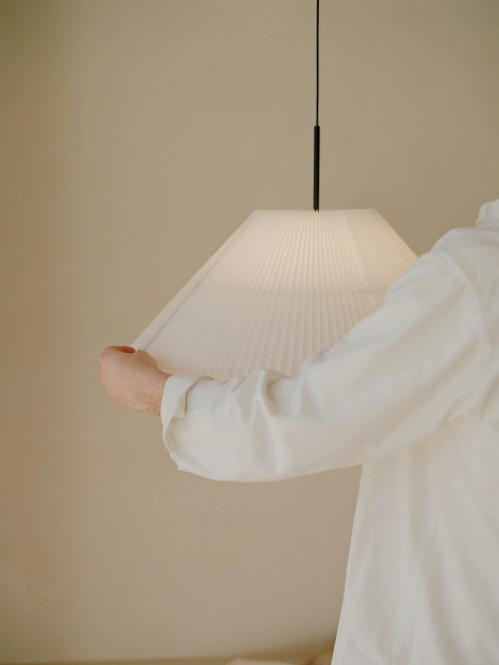 New Works Nebra Pendant Lamp, a modern Scandinavian pendant light featuring a wide, conical white pleated paper shade suspended from a sleek black cord. This minimalist ceiling lamp casts a soft, diffused glow, perfect for creating a warm ambiance in living rooms, dining areas, or bedrooms with a Japandi or contemporary design aesthetic.