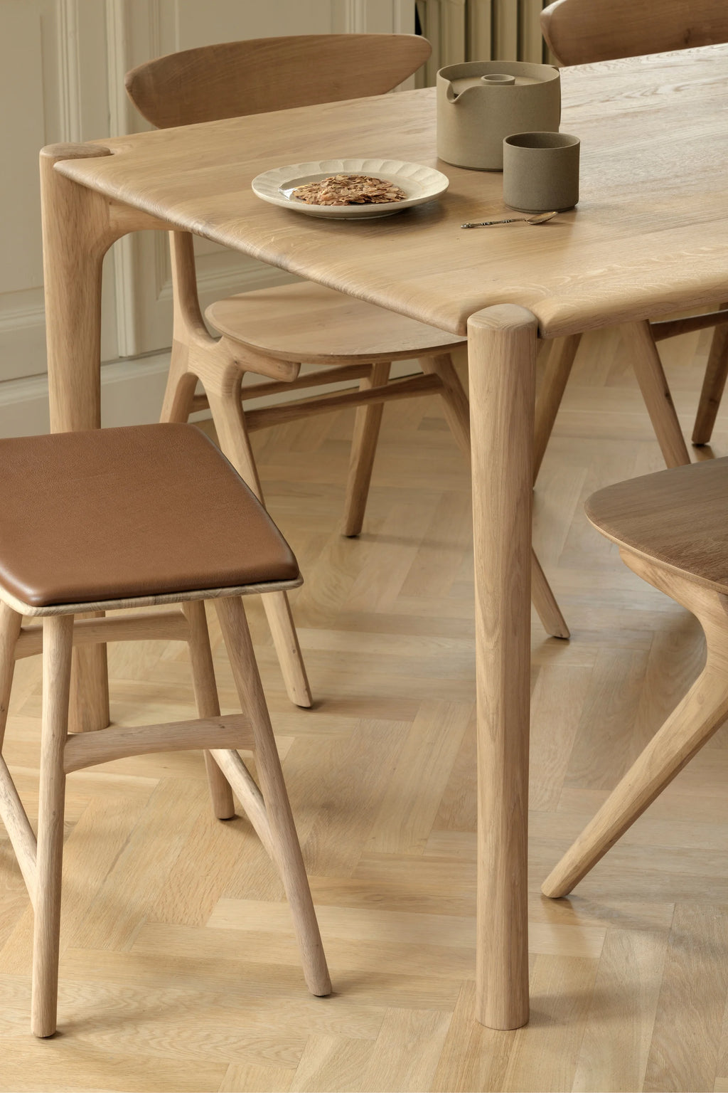 Ethnicraft PI Dining Table, a modern minimalist dining table crafted from solid natural light oak wood with a smooth, visible grain and distinctive rounded edges. Its substantial, organically shaped legs seamlessly integrate into the tabletop, creating a fluid Scandinavian design. Perfect as a centerpiece for contemporary dining rooms or open-plan living spaces.