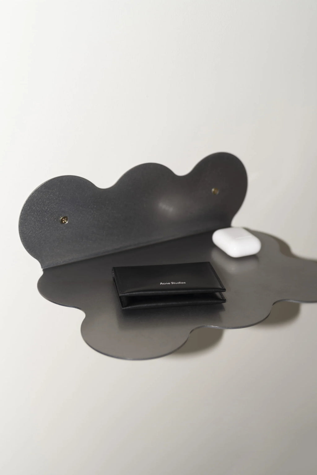 NG DESIGN's "Object 095 Shelf" in polished dark gray steel, with a distinct cloud-like wavy edge design mounted with gold screws, holding a black card holder and white earbuds on a white background. Creates a modern, minimalist display.